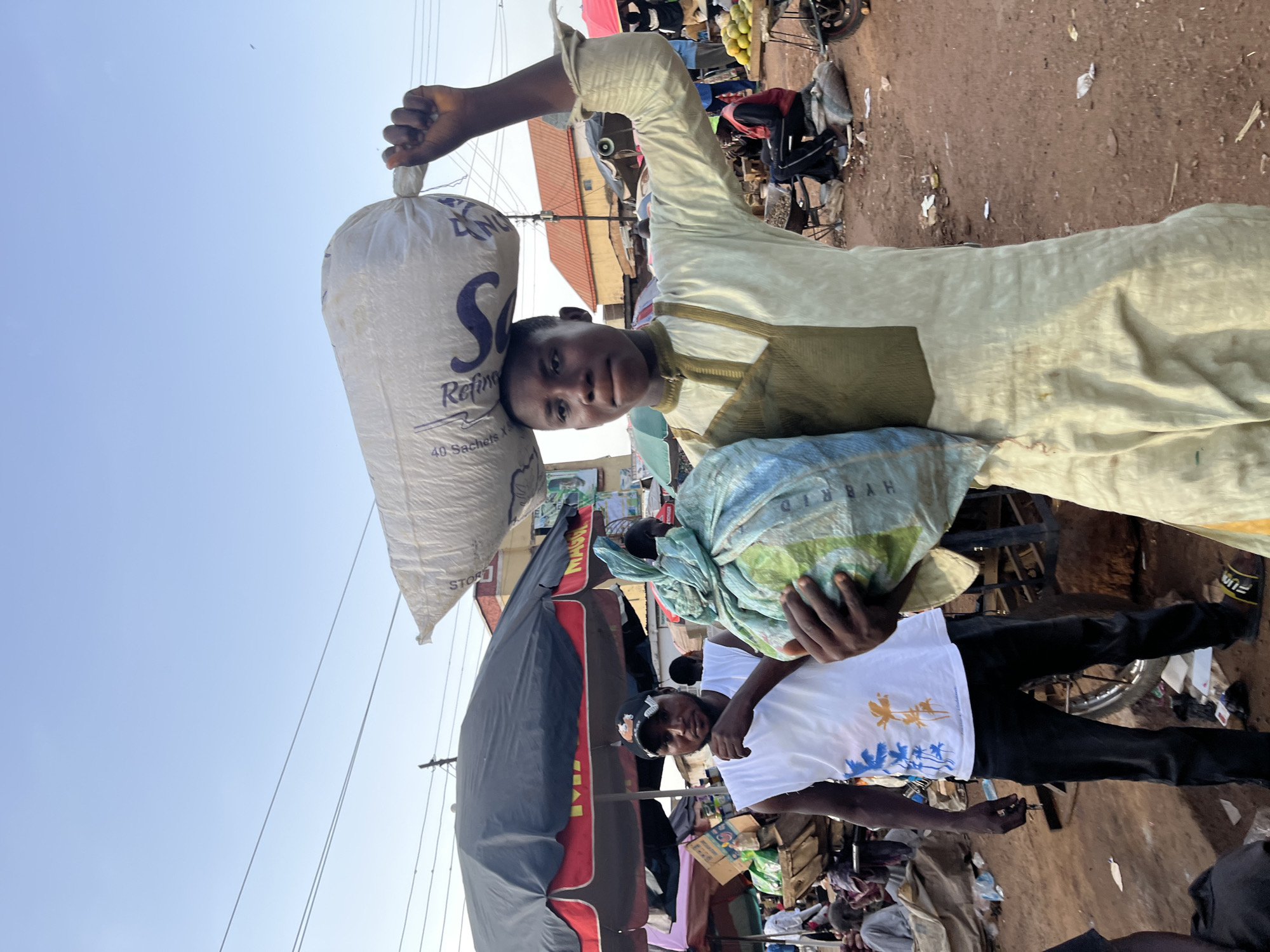 A young Black male, wearing a pale yellow traditional garment, carries a large, white sack of goods balanced on his head. He is in the foreground, facing slightly left, holding another large, blue-wrapped bundle of goods in his hands. To his left, another Black male, wearing a white sleeveless shirt and a black baseball cap, stands with his hand near his chin. Behind them, a bustling outdoor market scene unfolds. Various stalls with umbrellas and awnings display goods. People are visible in the background, some sitting and some standing. The ground appears to be dirt, with scattered debris. The sky above is clear and pale blue. The overall impression is a vibrant market in Rafin Par, Nigeria.