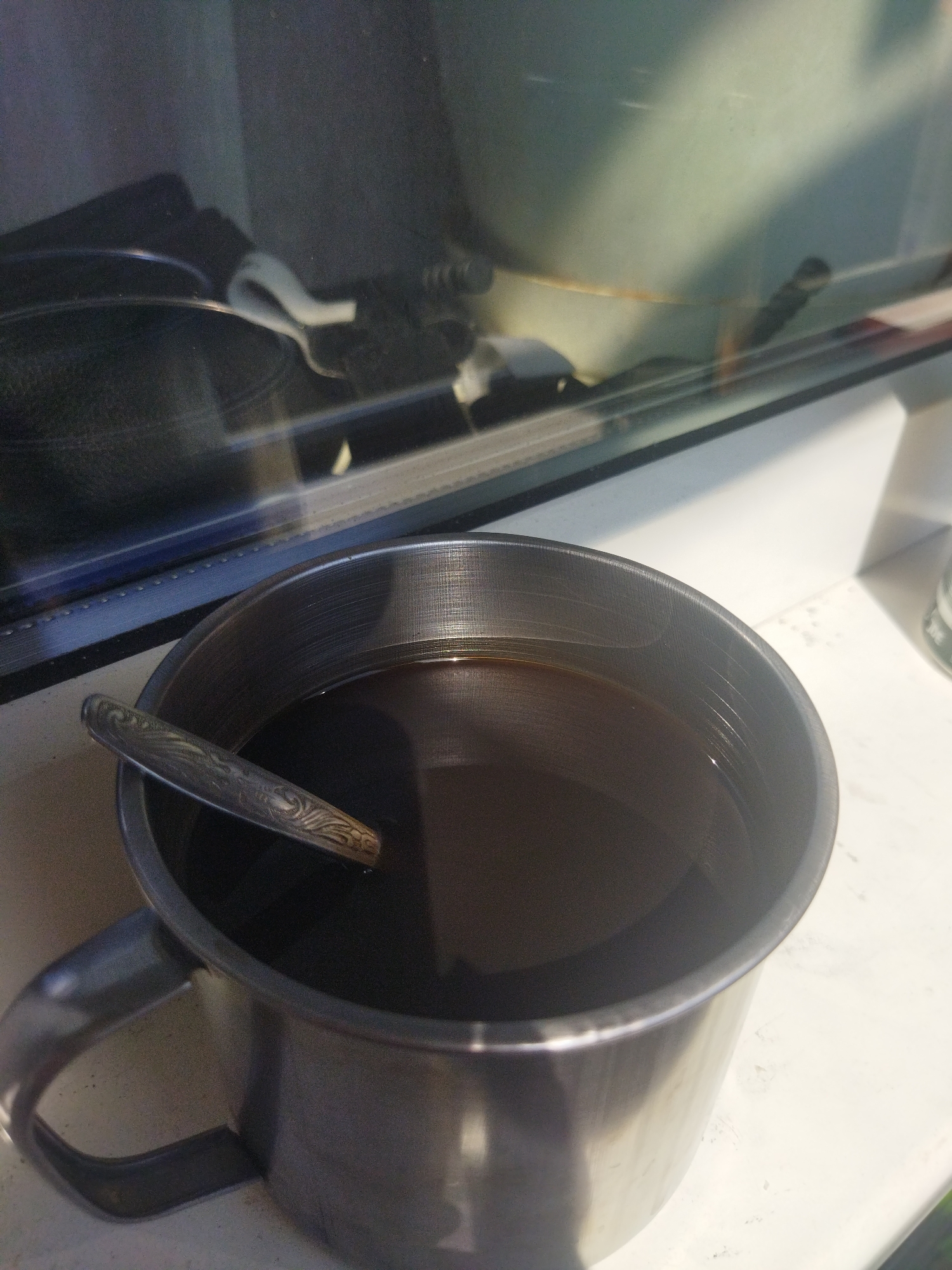 A close-up shot shows a metal mug filled with dark liquid, likely coffee or tea, and a spoon inside, placed on a light-colored surface near a window. The window reflects some items, possibly a dark bag, and a greenish object outside. The location is Kramatorsk, Ukraine. No people are directly visible in the image. The primary objects are the metal mug, liquid, spoon, and window. No specific actions or interactions are depicted other than the mug sitting on the surface. The environment appears to be indoors.