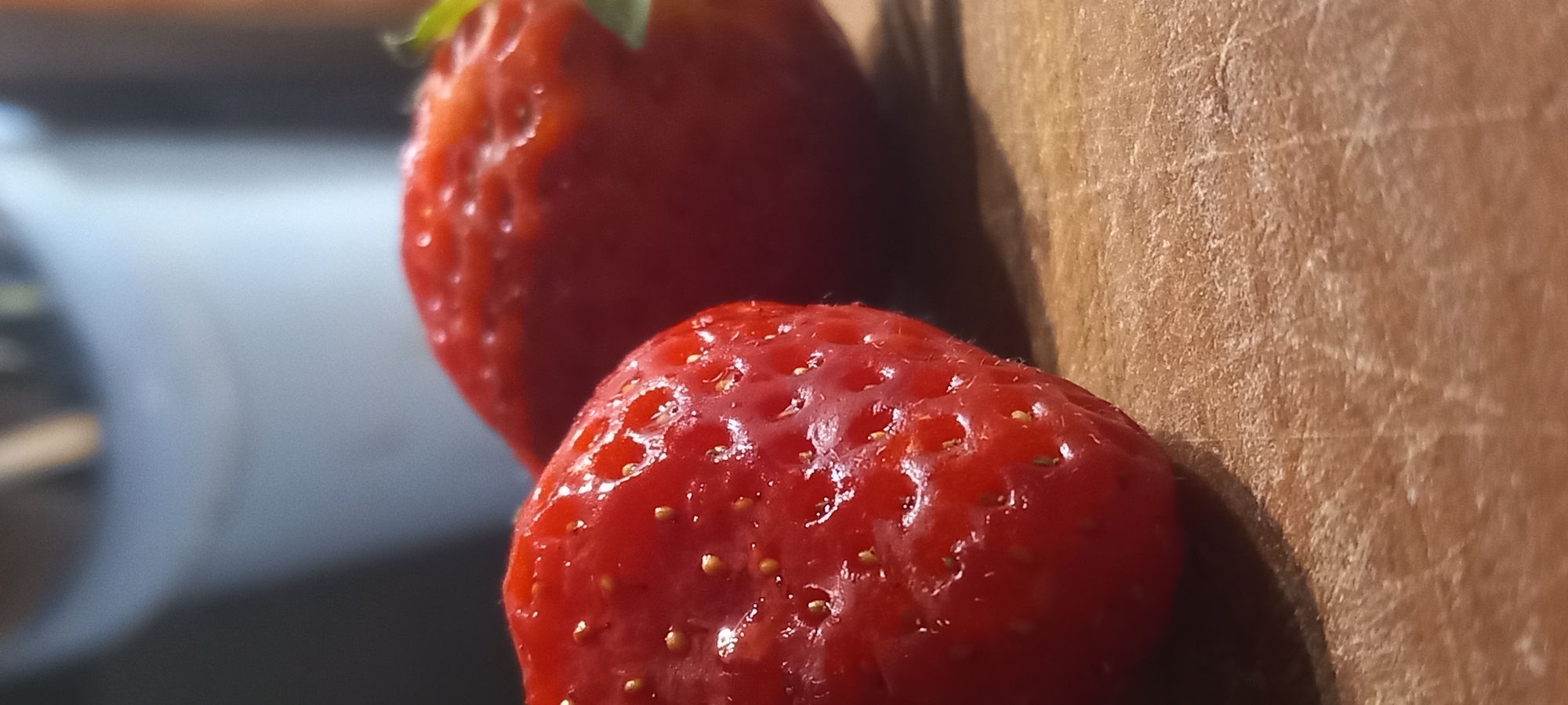 The image displays two ripe strawberries on a wooden surface. The strawberry in the foreground is sharply in focus, revealing its red skin covered in small, yellow seeds and a textured surface. The second strawberry, positioned slightly behind and to the right, is softer in focus, but also appears red and healthy. The wooden surface has a visible grain and appears slightly worn. The background is blurred, suggesting it is out of focus, with hints of a light-colored object and darker tones, possibly indicating a kitchen or indoor setting. There are no people or animals present, and no discernible landmarks or context to place the scene specifically within Kryvyi Rih, Ukraine.