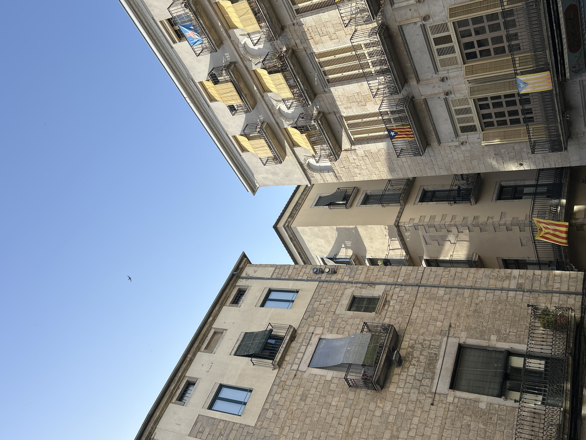 This is a low-angle shot of buildings in Girona, Spain, featuring a clear blue sky with a distant airplane. The architecture is characterized by stone facades and balconies adorned with yellow awnings and flags. On the right building, balconies on multiple levels display yellow awnings, and two balconies prominently feature Catalan flags. The left building, constructed with rough-hewn stone, also has balconies, one of which is covered by a translucent tarp. Windows are visible throughout, with some shuttered and others reflecting the sky. The scene depicts a section of a historic urban environment.