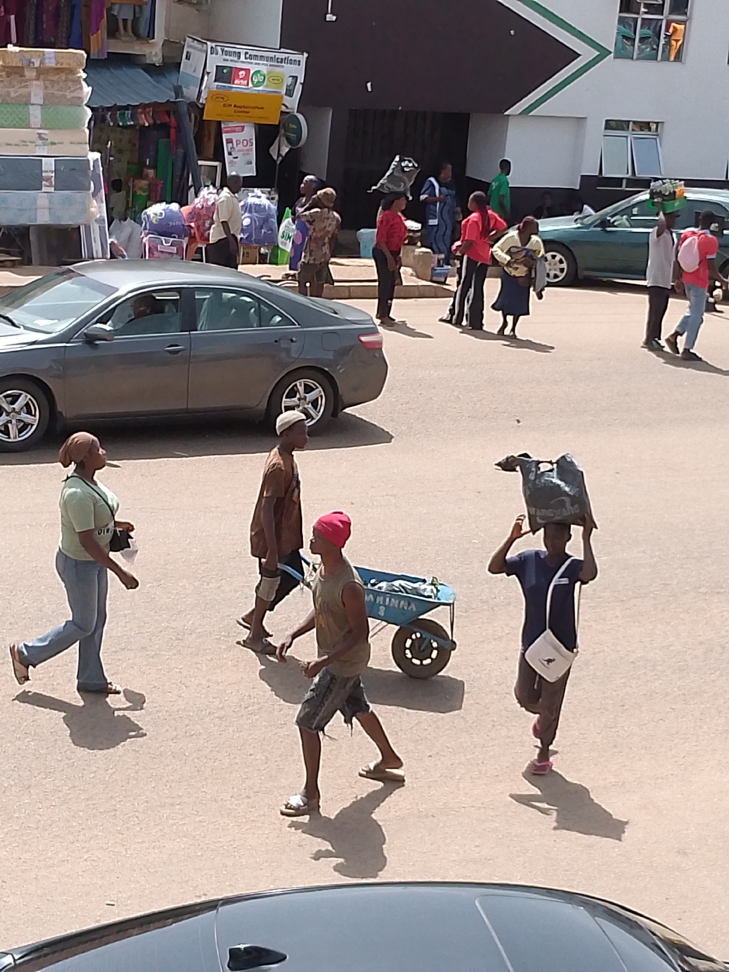 The image depicts a street scene in Bukuru, Nigeria, on a bright, sunny day, suggesting midday. The setting appears to be a bustling commercial area with shops lining the background. People are engaged in various activities, mostly walking or carrying goods.
In the foreground, a woman wearing a green shirt and blue jeans walks from left to right, carrying a black bag over her shoulder. Next to her, a man in a brown t-shirt and shorts walks past, wearing a light-colored cap. Behind them, another person, possibly a man, is seated next to a blue wheelbarrow.
Further back, a person in a blue shirt and dark pants carries a large black bag balanced on their head, with a white crossbody bag. To the left, a dark-colored sedan is parked, with a person visible inside. In the distance, other individuals are seen walking, some carrying shopping bags or other items. One person is carrying a basket of items on their head. A green car is also visible in the background.
The shops in the background display a variety of goods, including mattresses stacked high on the left. Signs are visible above the shops, one of which reads "DG Young Communications" along with logos for telecom providers like Airtel and Glo. Another sign displays "POS." The overall atmosphere suggests an active marketplace. The time of day is indicated by the strong shadows cast by the people and objects.
