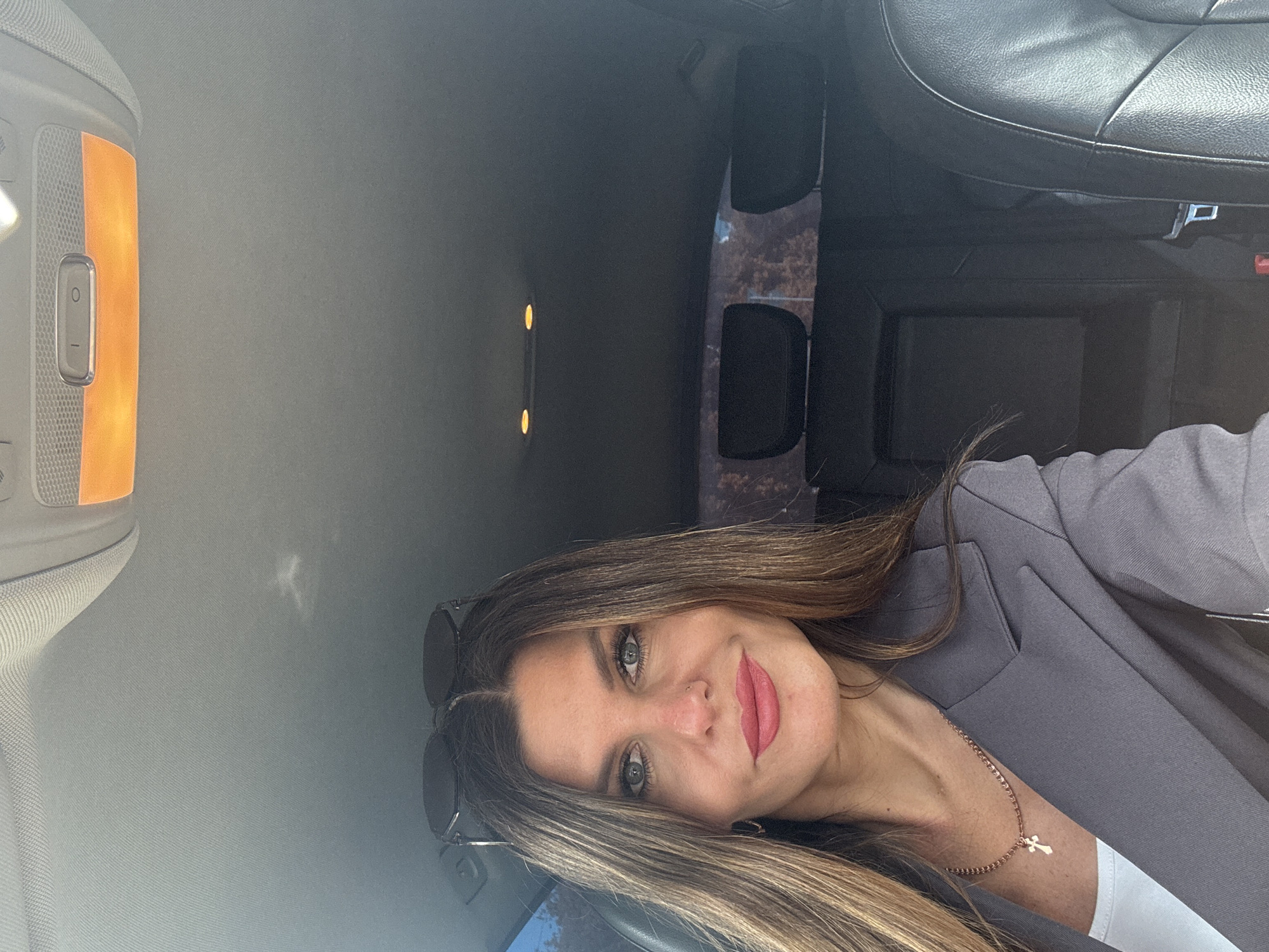 A woman with medium-length, highlighted brown hair and light eyes is seated in what appears to be the back of a vehicle. She is wearing a gray blazer over a white top and a delicate gold chain necklace with a cross pendant. A pair of sunglasses is perched on her hair. The interior of the vehicle is visible, including black leather seats, interior lighting, and portions of the ceiling. The location is not discernable as the visible surroundings offer no information to assist in identifying its location.