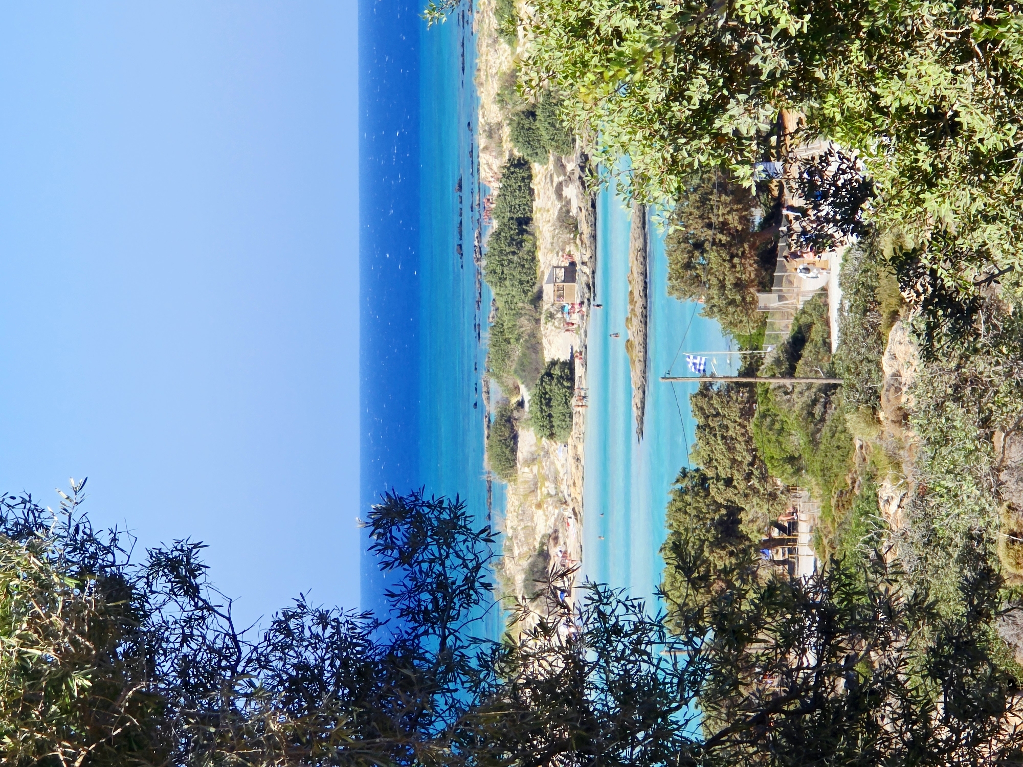 This is an eye-level, medium shot of a scenic coastal area in Greece, likely Elafonisi Beach, Crete, due to the characteristic pink sand and turquoise waters. In the foreground, dark green, leafy branches of trees frame the view. Beyond the trees, a bright blue sky meets the deeper blue of the sea. The middle ground features a sandy beach dotted with scattered rocks and low green shrubs. Several small groups of people are visible on the beach and in the shallow, clear turquoise water. Some appear to be sunbathing or walking, while others are swimming. Small structures, possibly beach huts or changing rooms, are present on the sand. A Greek flag flies from a pole on the right side of the image. The background is dominated by the expansive blue ocean.