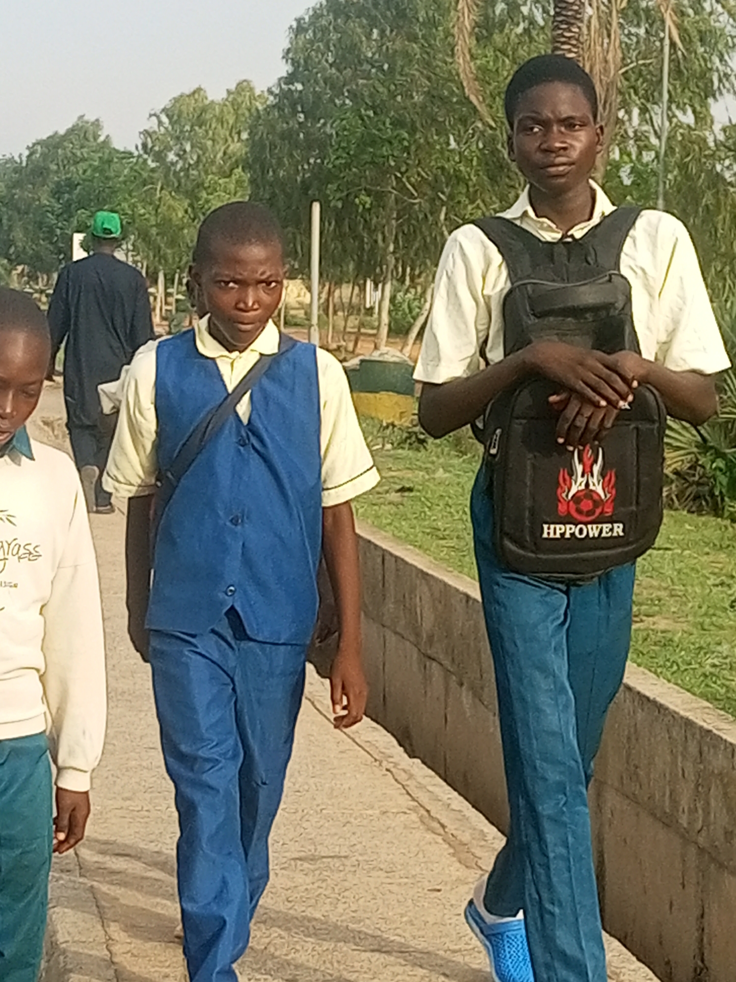 In Gombe, Nigeria, two young boys are walking down a pathway. The boy on the right is wearing a light yellow collared shirt, blue trousers, and a black backpack with the logo "HPPOWER" visible. He has his hands crossed over the backpack. The boy on the left is wearing a yellow collared shirt and a blue vest, also with blue trousers. A third individual, partially visible, is walking ahead of them and wearing a green cap and a dark garment. The pathway appears to be made of concrete or stone, bordered by a low concrete wall. Behind the pathway, there is lush green vegetation and trees, indicative of an outdoor environment. The sky is bright and clear, suggesting daytime.