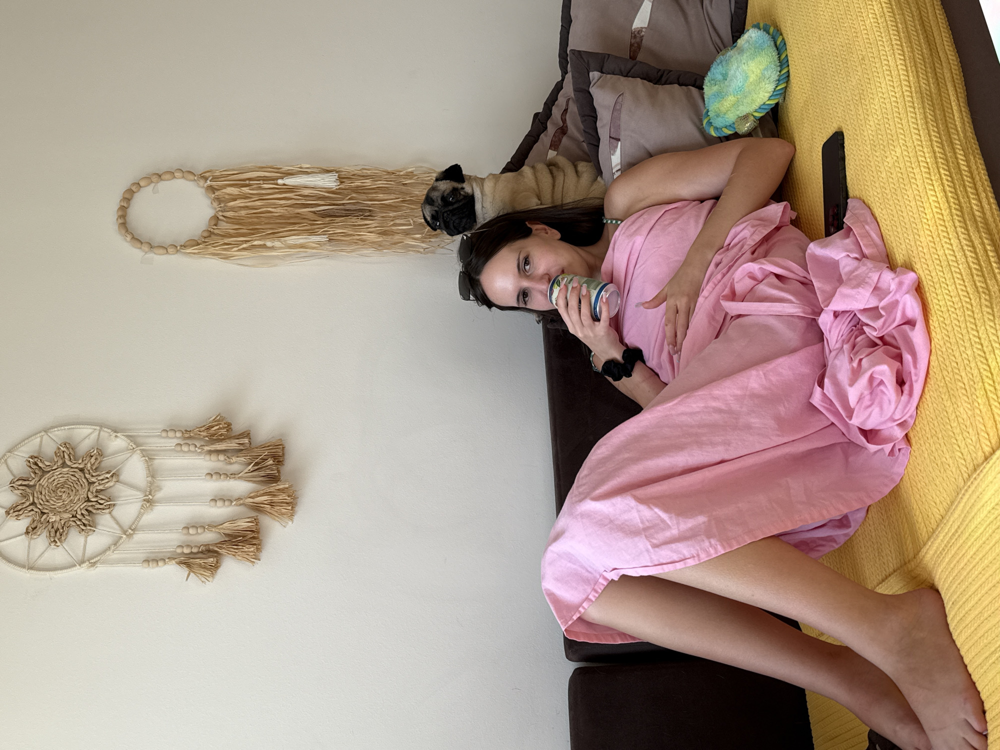 A woman lies on a sofa, holding a can to her lips. She is wearing a pink dress and has sunglasses perched on her nose. A pug is lying next to her on the sofa, appearing to be asleep. The sofa is covered with a yellow blanket. The scene appears to be indoors, with a plain wall in the background adorned with two macrame wall hangings. The lighting suggests it is daytime. The woman's expression is neutral. There is no visible text in the image.