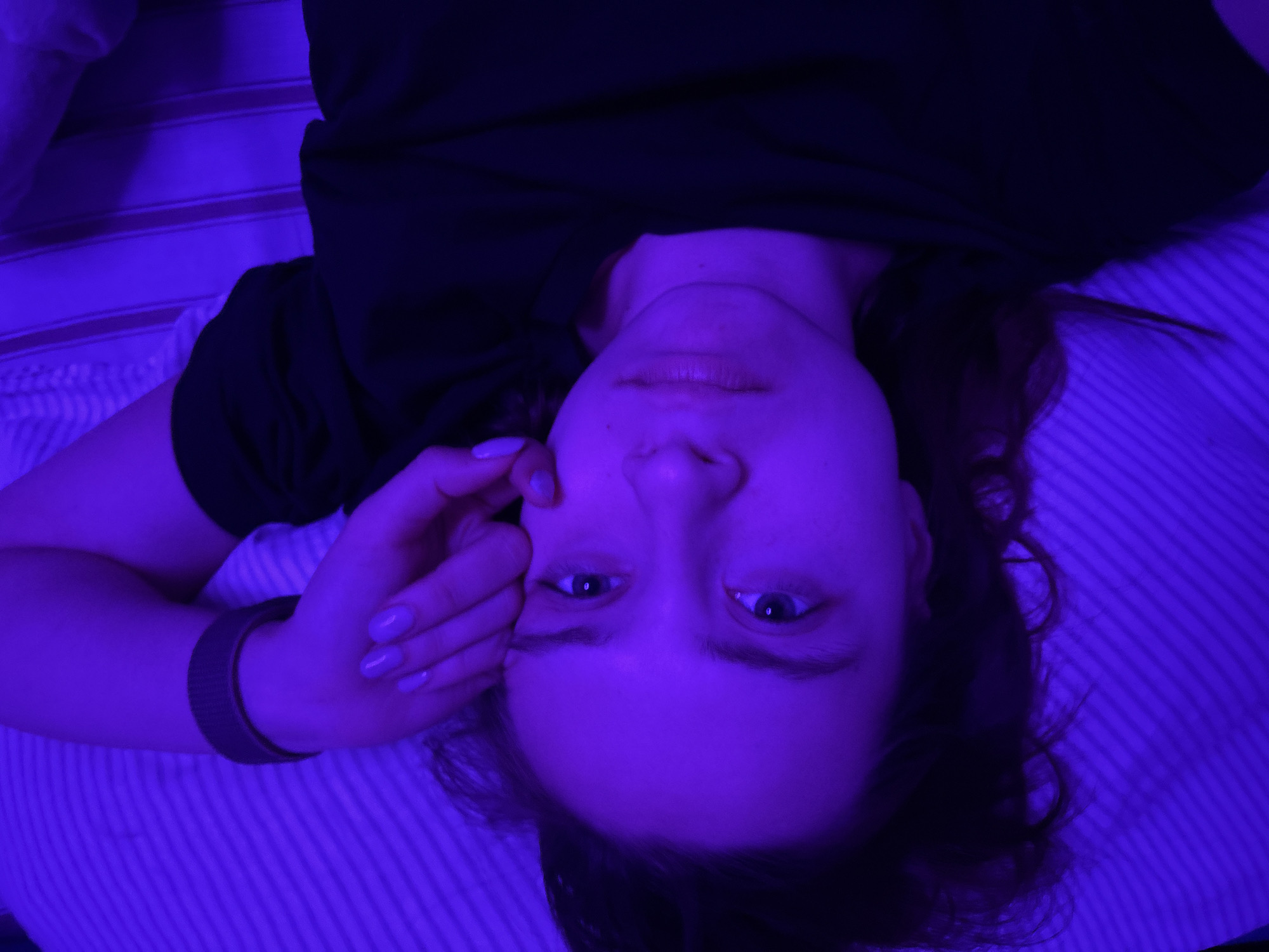 A high-angle, medium close-up shot shows a young adult female lying on her back in an indoor setting, looking directly at the camera. She has dark hair and is wearing a black t-shirt. Her left hand, adorned with light purple nail polish, is gently resting near her temple. A dark, possibly fabric, wristband is visible on her left wrist.
The entire scene is illuminated by striking artificial purple lighting, which bathes her skin, clothing, and the striped bedding (likely a pillow or sheet) in a deep violet hue. The stripes on the bedding run diagonally and horizontally across the frame. Her facial expression appears neutral and direct, with her eyes wide open. The purple illumination suggests either a nighttime setting or a deliberately darkened room with specialized lighting.
There is no visible text in the image. The location context provided for this image is Прикамский, Russia.