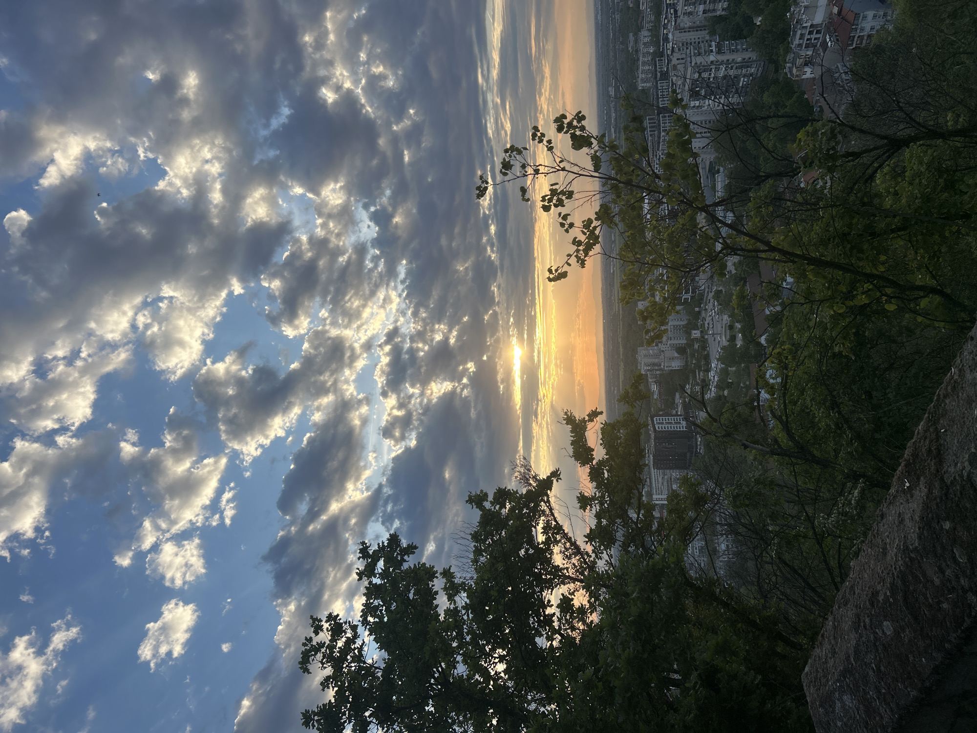 The image shows a view of Plovdiv, Bulgaria, at sunset, likely from a higher elevation point. The sky is filled with clouds illuminated by the setting sun. The sun's rays are visible, creating a bright streak in the sky. Below the horizon line, a cityscape can be seen, suggesting a densely populated urban area. In the foreground, there are trees and foliage, partially obscuring the view of the city. The angle of the shot indicates that the photographer is likely standing on a hill or elevated platform overlooking the city. The colors are muted due to the sunset lighting, with oranges, yellows, and grays dominating the scene. The image suggests a blend of natural and urban environments.