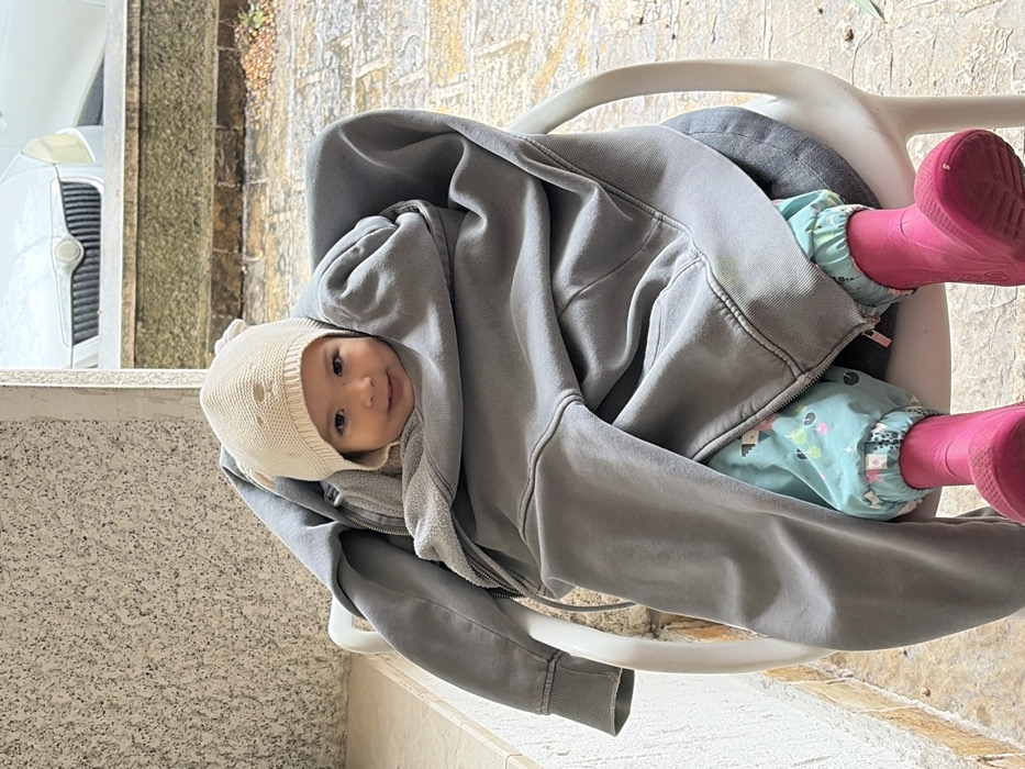 A young child is seated in a white plastic chair, positioned centrally. The child wears a light beige knitted hat with small ear-like protrusions, light blue patterned pants, and bright pink rubber boots. An oversized, dark grey zippered hoodie is wrapped around the child, largely obscuring the body, with only the head, lower legs, and boots visible. The child's face is smiling and directed towards the viewer. The immediate background to the left features a textured, light-colored aggregate wall. The ground beneath the chair and extending into the mid-ground consists of light brown or greyish flagstones. In the upper right background, the front portion of a light-colored vehicle, consistent with a white Skoda, is visible above a low stone wall or planter. The specific location within Budva, Montenegro, is not discernible from the visual evidence.