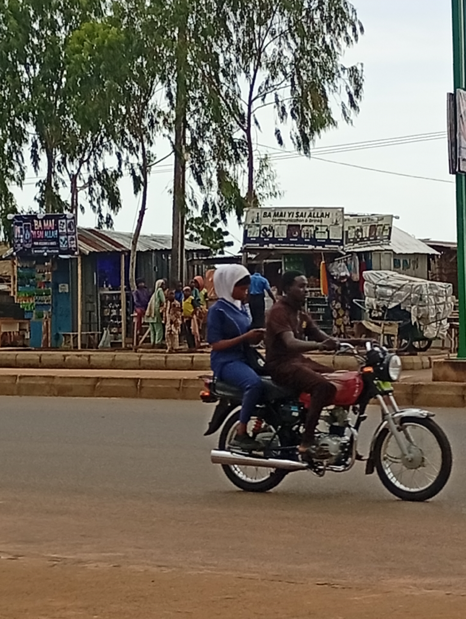 In Wuro Dole, Nigeria, a man and a woman are riding on a red motorcycle. The man is driving, wearing a brown shirt and pants. The woman is in the back, wearing a blue top and pants with a white hijab. They are on a paved road with shops and trees in the background. Several other people are visible in front of the shops, some walking and some standing. The shops appear to be small, open-air businesses. The overall scene depicts a busy street in a Nigerian town.