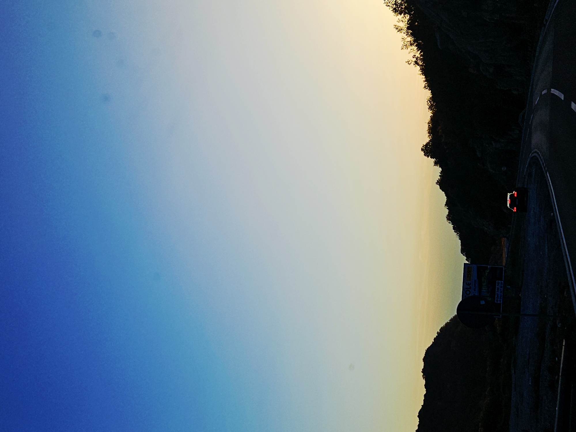 The image captures a scenic view of a winding road through a mountainous landscape at dusk. The sky transitions from a deep blue at the top to a pale yellow-orange near the horizon, suggesting either sunrise or sunset. Silhouetted against the sky are dark, rugged hills and trees, creating a dramatic contrast. A single red car is visible driving on the road, its taillights illuminated. In the foreground, the road is paved with white lane markings. There is a signpost with some text and an image on it located on the left side of the road, partially obscured by the car. The overall impression is one of a tranquil, remote setting.