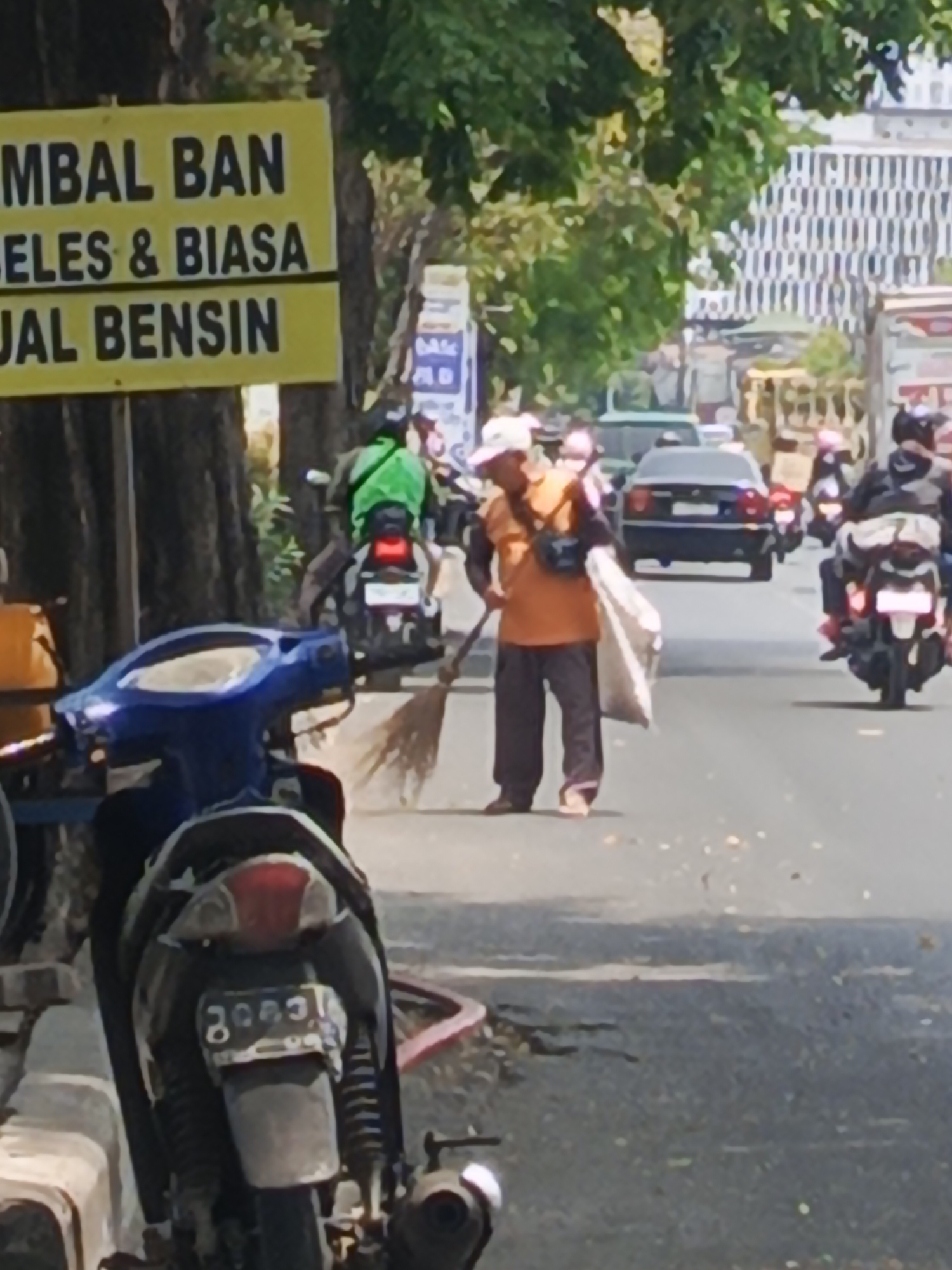 This is a medium-shot, eye-level, outdoor photograph taken on a sunny day on a street in Surabaya, Indonesia. The street appears to be a busy thoroughfare with traffic. In the foreground, a blue scooter is parked on the left side of the road. Further down the street, a person dressed in an orange shirt and dark pants is sweeping the road with a broom. They are carrying a large white bag over their shoulder. Behind the sweeper, there is a black car and several motorcycles, some with riders wearing helmets. In the background, there are trees, buildings, and a yellow sign with black lettering in Indonesian that reads "GUMBAL BAN SELES & BIASA JUAL BENSIN" (which translates to "Tire Repair, Used & Regular, Gasoline for Sale"). The overall impression is a snapshot of daily life on a bustling street in an Indonesian city.