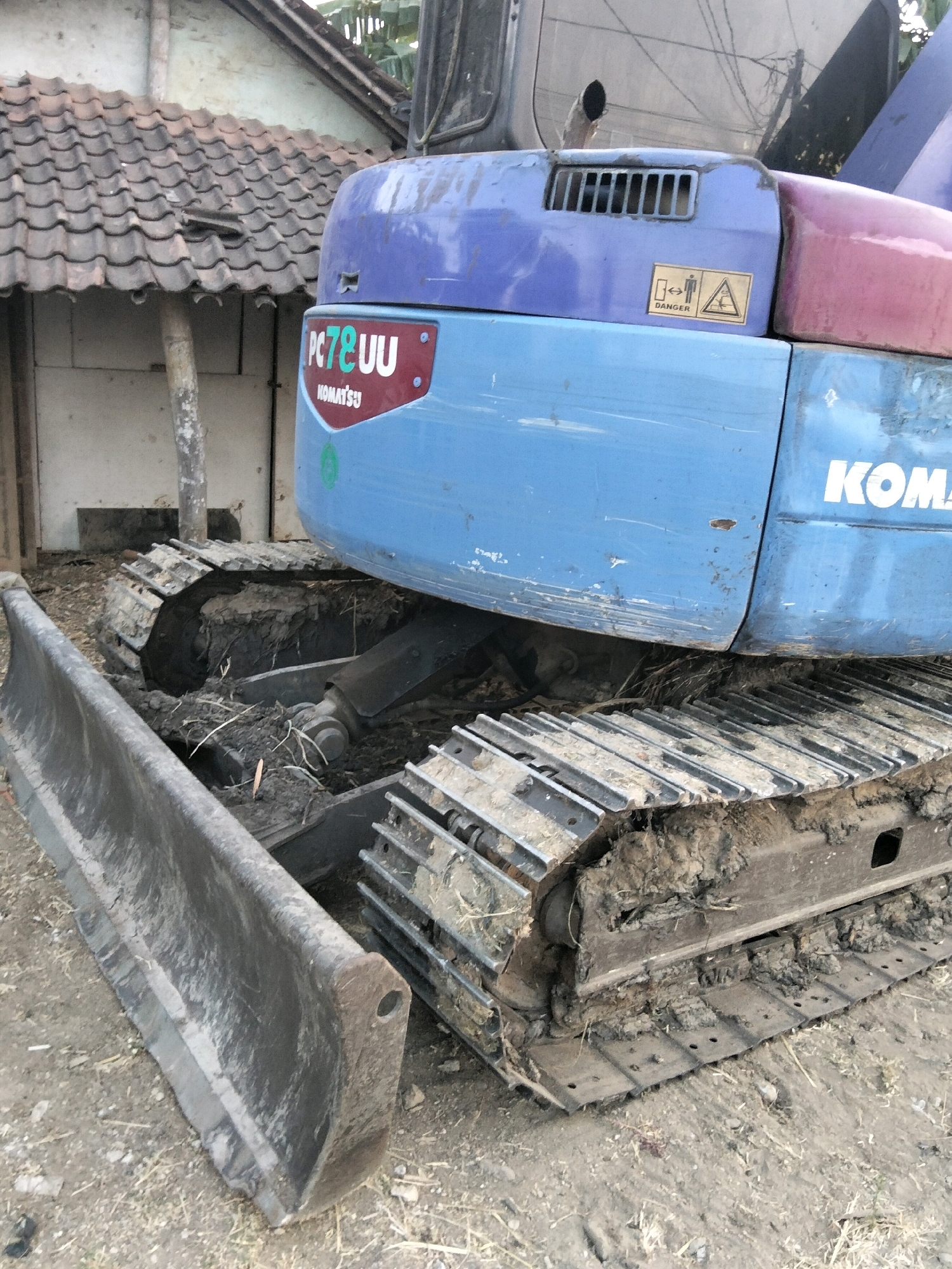 A Komatsu excavator with a blade is parked on a dirt ground outside a building. The building has a tiled roof and is partially visible in the background. The excavator is blue and red, with tracks covered in mud. A "PC78UU" decal is visible on the side of the excavator. There is also a yellow "DANGER" sticker on the excavator. The image appears to have been taken in Mrican, Indonesia.