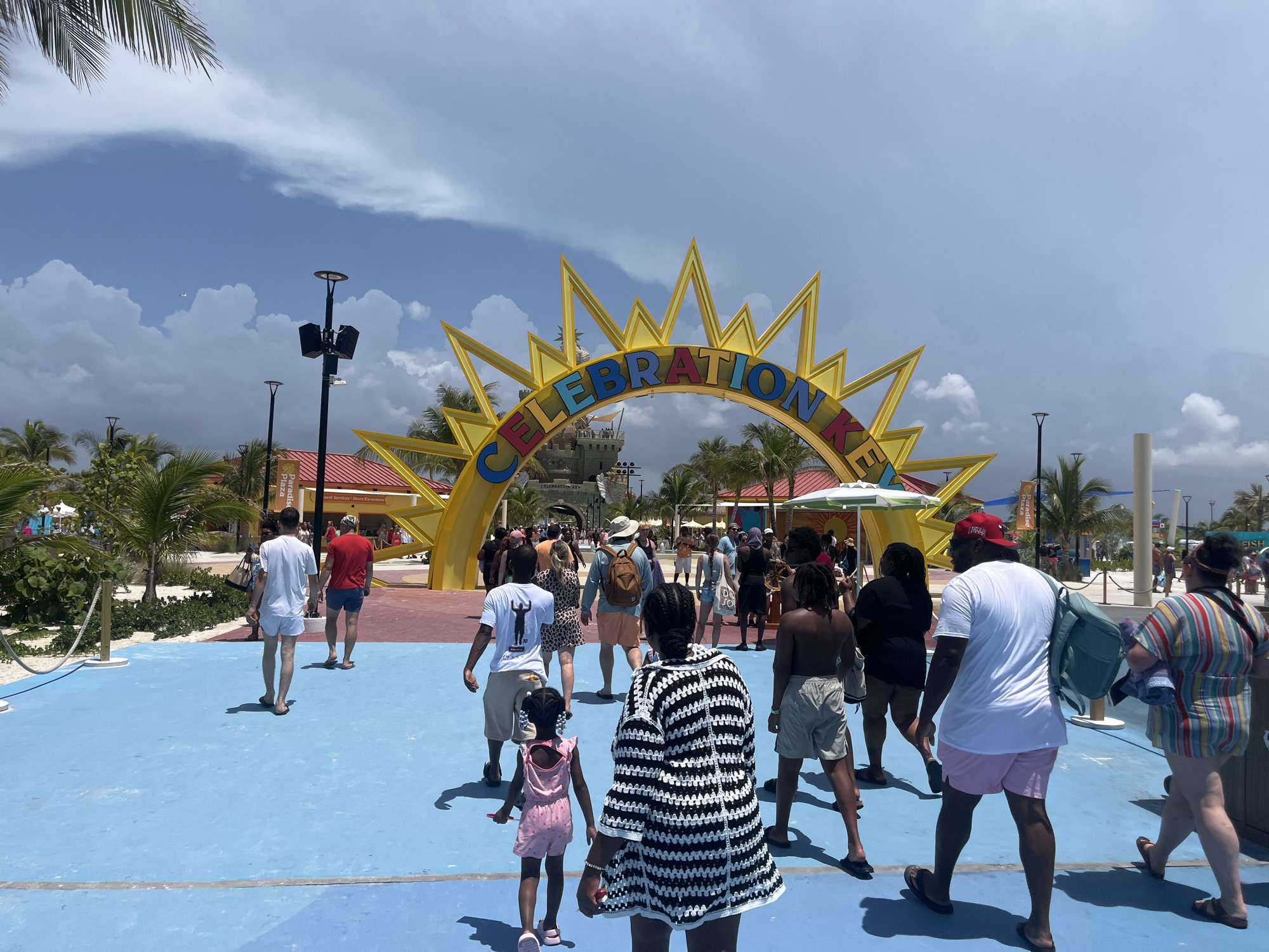 An overhead shot captures a group of people walking on a bright blue pavement towards a large, yellow sun-shaped archway with the word "CELEBRATION" in colorful block letters. The archway is adorned with lights and leads to a castle-like structure. Palm trees and a few buildings with red roofs are visible in the background, under a partly cloudy sky. The scene appears to be a public space, possibly a theme park or a cruise port, given the festive atmosphere and attire of the people. This location is consistent with a part of City of Freeport, Bahamas, known for its tourist attractions and cruise terminals.