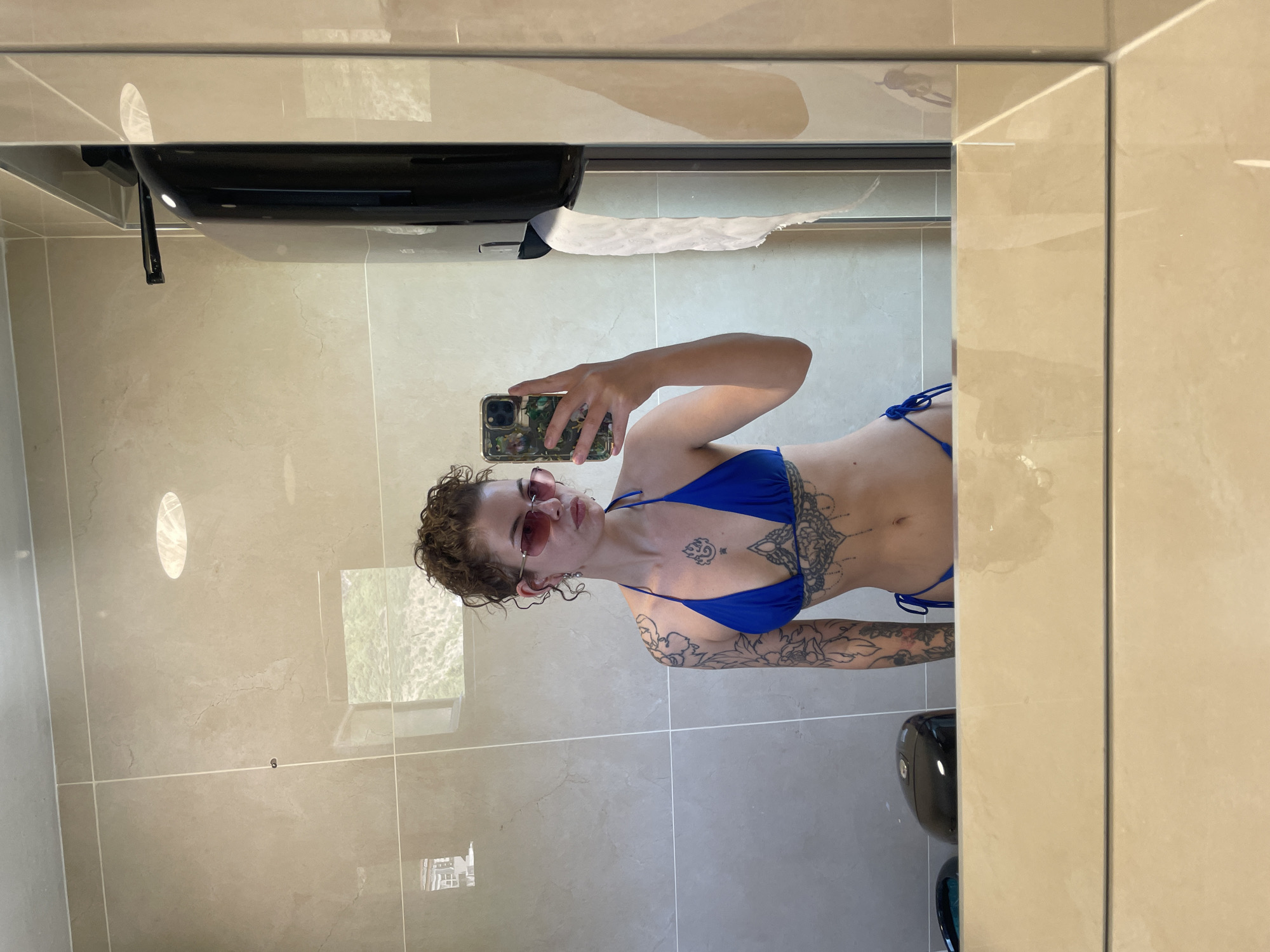 A young woman with curly brown hair and pink sunglasses stands in front of a tiled wall and takes a mirror selfie. She is wearing a blue bikini and has tattoos on her arms and torso. A black dispenser hangs on the right side of the wall, and a black appliance is visible on the left. The scene appears to be a restroom in a hotel or resort located in Bečići, Montenegro.