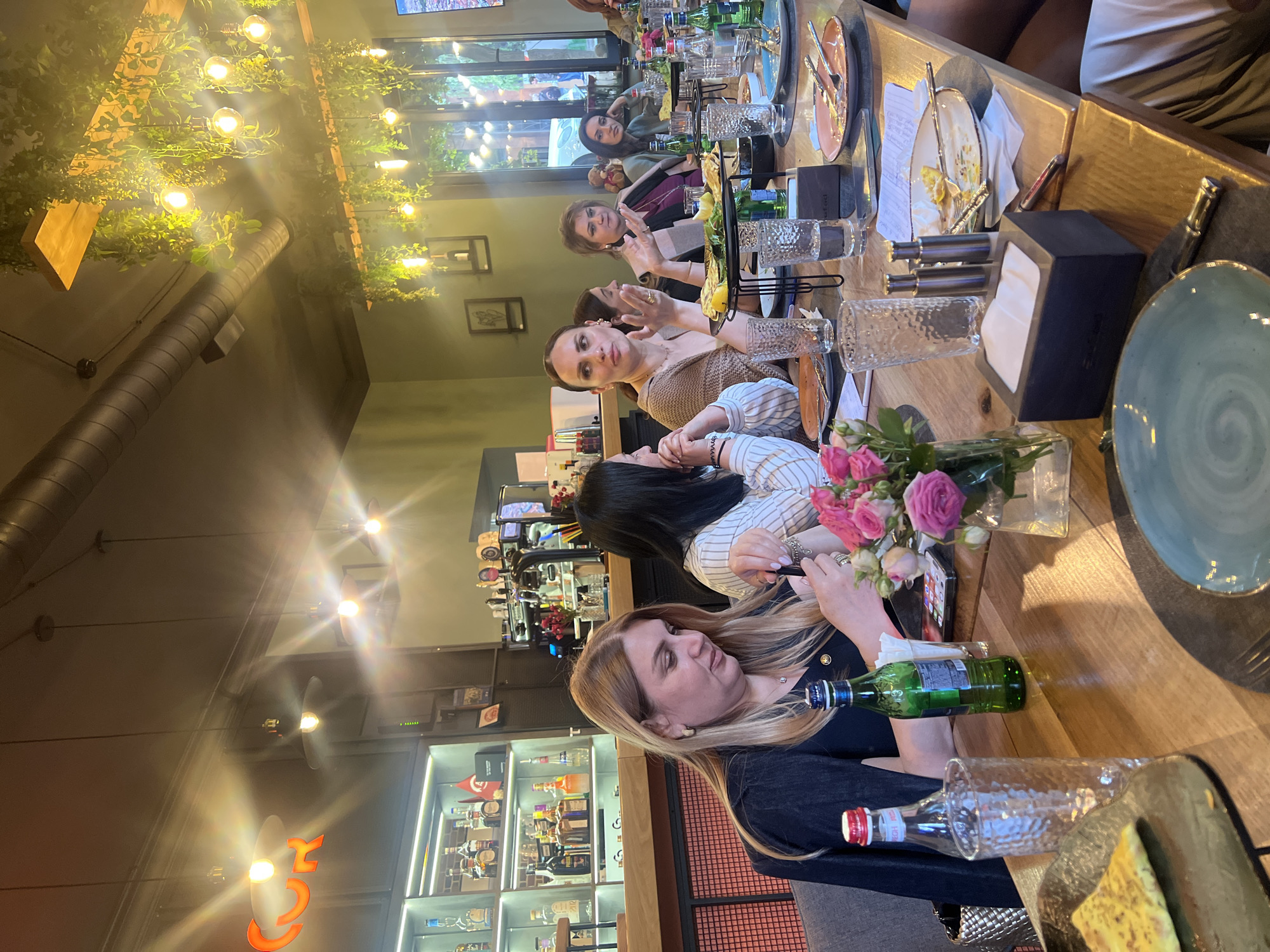 The image shows a group of six women seated at a long wooden table in what appears to be a restaurant. The table is set with plates, glassware, bottles of water, and a bouquet of pink roses in a vase. The women are dressed in casual attire. The background features a display case with various bottles of liquor and soft drinks, indicative of a bar area. Hanging greenery with lights is visible above the table. The location is likely in Yerevan, Armenia, based on the restaurant's decor and overall ambiance.