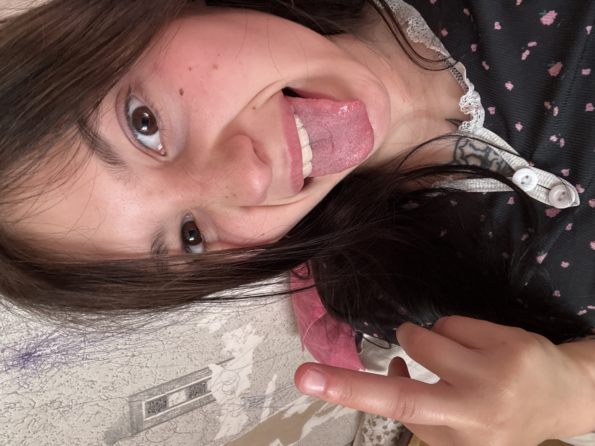 A close-up, rotated image captures a young person with long, dark brown hair and dark brown eyes, making a playful expression. Their head is oriented upwards and to the right from the viewer's perspective. The person is sticking out their tongue and smiling, revealing a mischievous or happy emotion. Their face shows several freckles or moles.
They are wearing a dark top or dress featuring a subtle pattern of small pink flowers and white lace trim along the collar or neckline. White buttons are visible on the front of the garment near the shoulder. A dark-colored tattoo is partially visible on the person's upper chest or shoulder, peeking out from under their clothing. One hand is visible in the lower right of the frame, with the index finger extended.
The background is a light-colored, textured surface, possibly a sheet or fabric, adorned with faint patterns, blue pen scribbles, and a stylized outline design resembling a building structure. Several loose strands of hair are scattered across this surface. The scene suggests an informal indoor setting, likely a home environment in Фоки, Russia. There is no visible text in the image.