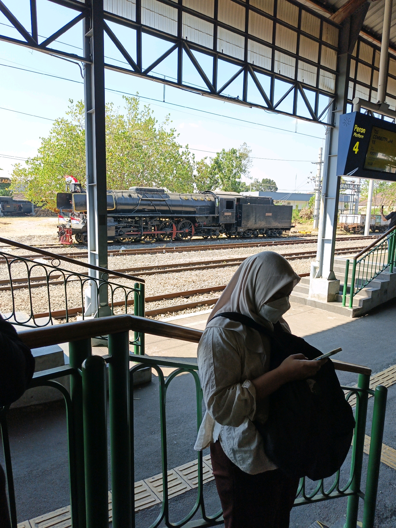 A wide shot captures a scene at the Surakarta Train Station in Surakarta, Indonesia, on a bright, sunny day. In the foreground, a person wearing a light-colored hijab and a light beige long-sleeved shirt, and dark red pants, is looking down at their mobile phone while holding a black backpack on their shoulder. They are positioned behind a green metal railing with a wooden top.
In the mid-ground, train tracks stretch across the frame. A vintage black steam locomotive, adorned with two small Indonesian flags on its front, is parked on the tracks. Beyond the locomotive, other train cars and carriages are visible, along with some trees.
In the background, a platform with a staircase is situated to the right, with a digital display board indicating "Peron 4" (Platform 4) and departure information for the "Commuter Line YK" to Yogyakarta at 14:00. The station's roof structure, made of metal beams and translucent panels, covers the platform area. The sky above is clear and blue.
