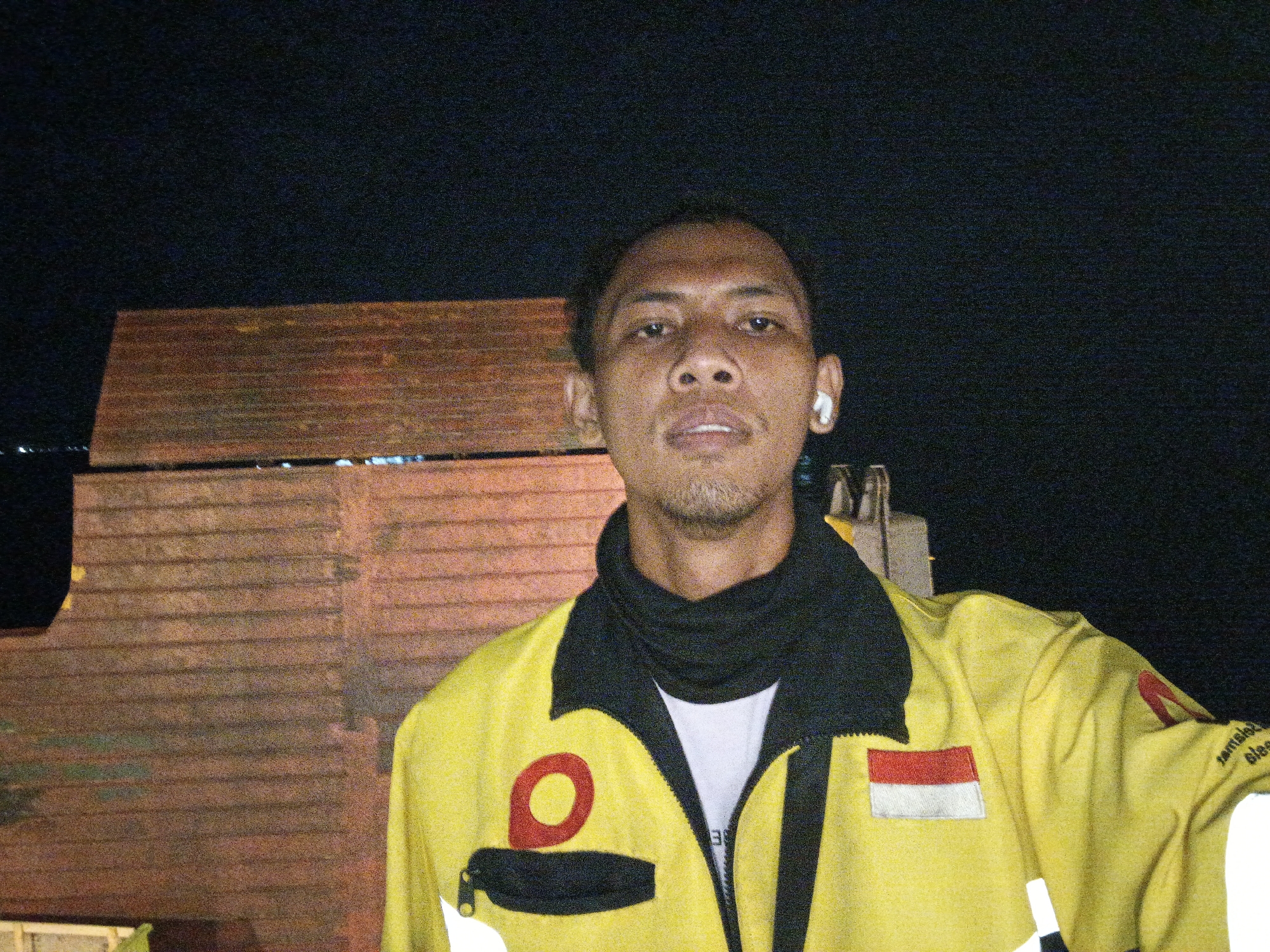 In a dimly lit outdoor setting, a man stands against a dark background with a wooden structure behind him. He is wearing a bright yellow jacket with black trim, a white t-shirt underneath, and a black neck gaiter. The jacket features a red circular logo on the left chest and a rectangular patch with red and white horizontal stripes on the right chest. The man has a light beard and is wearing a white wireless earbud in his right ear. The overall scene suggests a nighttime setting in Banyuwangi, Indonesia, possibly near a transportation hub or industrial area, given the industrial-looking wooden structure.