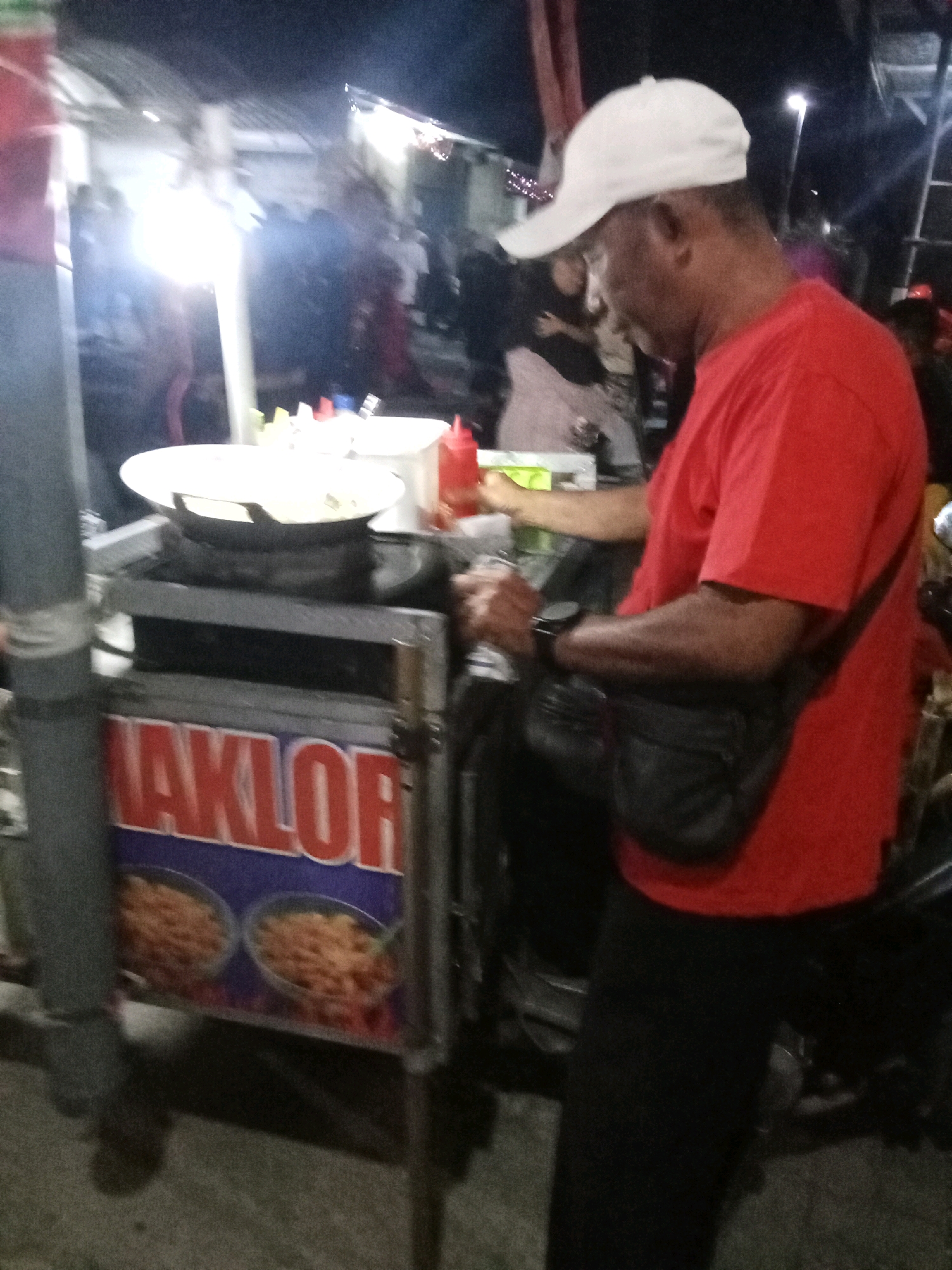 At night in Keplaksari, Indonesia, a man in a red t-shirt and white baseball cap stands behind a food cart. The cart is decorated with an advertisement that reads "MAK LOR" and displays images of two bowls of food. The man is wearing a black fanny pack around his waist and appears to be interacting with the food cart, possibly preparing food or attending to a customer. In the background, other people are visible, suggesting a lively street scene. The lighting is dim, typical of nighttime street vending.
