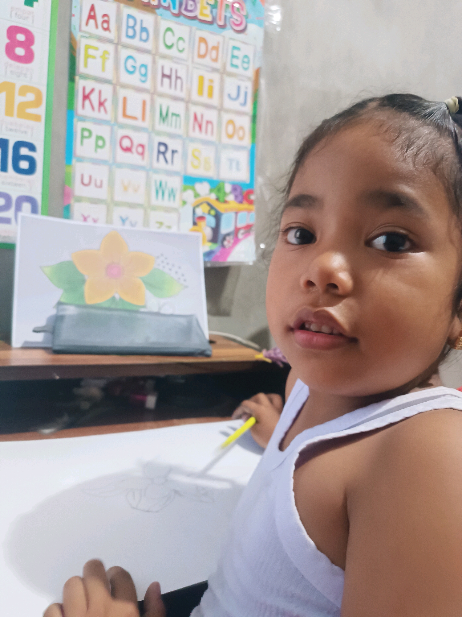 A young girl with dark hair tied in a ponytail and wearing a white tank top is seated at a table, looking towards the camera. She is holding a yellow pencil and appears to be drawing on a white sheet of paper. In the background, an alphabet chart and a picture of a yellow flower with green leaves are visible. The scene is indoors, likely in a home environment in Tabanan, Indonesia, given the context.