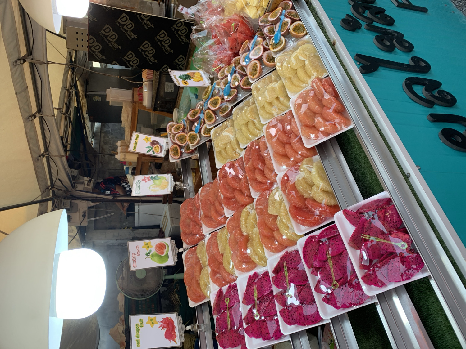 This image depicts a vibrant fruit stall, likely situated in a market in บ้านบางรักษ์, Thailand. The stall is brimming with an assortment of pre-cut and pre-packaged tropical fruits, meticulously arranged in white plastic trays and sealed with clear plastic wrap. A prominent feature of the display is the abundance of red and yellow pomelo segments, along with striking deep red dragon fruit slices. Numerous halved passion fruits, some accompanied by small blue plastic spoons, are also presented, ready for immediate consumption. Pre-cut pineapple slices are visible among the offerings.
Various signs identify the produce, with visible text including "red dragon fruit," "Siam Ruby Pomelo," "pomelo," and "passion fruit." One sign partially displays "100B," likely indicating a price in Thai Baht. The fruits are showcased on multi-tiered shelves, set against a backdrop of a partially visible alleyway or back area. Overhead, a canvas awning provides shelter, and white dome-shaped lamps provide artificial illumination. A fan is visible, suggesting a warm climate. In the background, several dark boards bear the repeated text "DD PLUS Film Faced Plywood." To the far right, a blue-green surface features bold black lettering or numbers, specifically "16." The scene portrays a well-organized and colorful market stall dedicated to selling fresh, ready-to-eat fruit. No people are clearly visible within the frame.