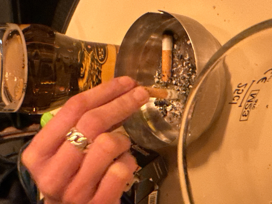 The image captures a close-up view of a person's hand holding a lit cigarette over a metallic ashtray, set in what appears to be an indoor environment. The hand, with light skin, is prominent in the foreground, holding the cigarette between the index and middle fingers. A silver-colored ring with a distinctive chain-link pattern is visible on the ring finger of the hand.
The lit cigarette has a visible cherry and a small amount of ash at its tip, which is being held directly above a round, silver-colored metal ashtray. Inside the ashtray, there's a significant amount of grey ash and at least one other orange-filtered cigarette butt resting in one of the ashtray's notches.
To the left of the hand and ashtray, a glass or mug filled with a dark liquid, possibly beer, is partially visible. The glass has a label with dark and yellowish tones and some intricate design, possibly a crest or animal motif. The text "25cl" is clearly visible on the lower part of this glass, indicating its volume.
Below the ashtray and partially obscured by the hand, a dark-colored object, likely a cigarette pack, can be faintly seen.
To the right, a light-colored, possibly ceramic or stoneware object (such as a plate or a large coaster) is angled upwards, showing its underside or rim. Several pieces of text are visible on this object, oriented upside down relative to the main image composition: "CE", a rectangular box containing "WS3", and "52cl".
The overall lighting in the image is warm, with yellowish tones, suggesting an indoor setting, possibly during the evening or night. The scene conveys a casual and relaxed atmosphere, centered around the act of smoking and drinking.
