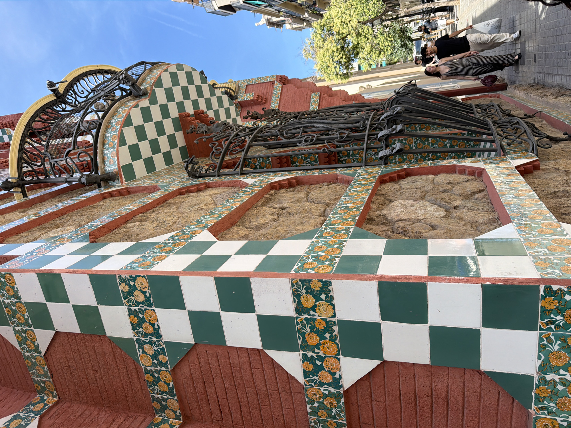 The image depicts the facade of Casa Vicens, a historic house located in the Gràcia district of Barcelona, Spain. The building's exterior features a vibrant mosaic of green and white checkered tiles, interspersed with decorative bands of floral tiles in shades of orange and yellow. The textured, reddish-brown brickwork is visible between the tiled sections. A distinctive wrought-iron balcony with intricate decorative elements is visible on the upper part of the building, also adorned with checkered and floral tiles. A large, ornate wrought-iron window grille is prominent on the facade. In the foreground, a sidewalk with patterned paving stones runs alongside the building. Two people are walking on the sidewalk: a woman in a patterned jumpsuit and a man in a black t-shirt and light-colored pants, holding a white plastic bag. Further down the street, other pedestrians and trees are visible, under a clear blue sky. The architectural style is characteristic of Antoni Gaudí's early work, reflecting Mudejar and oriental influences.