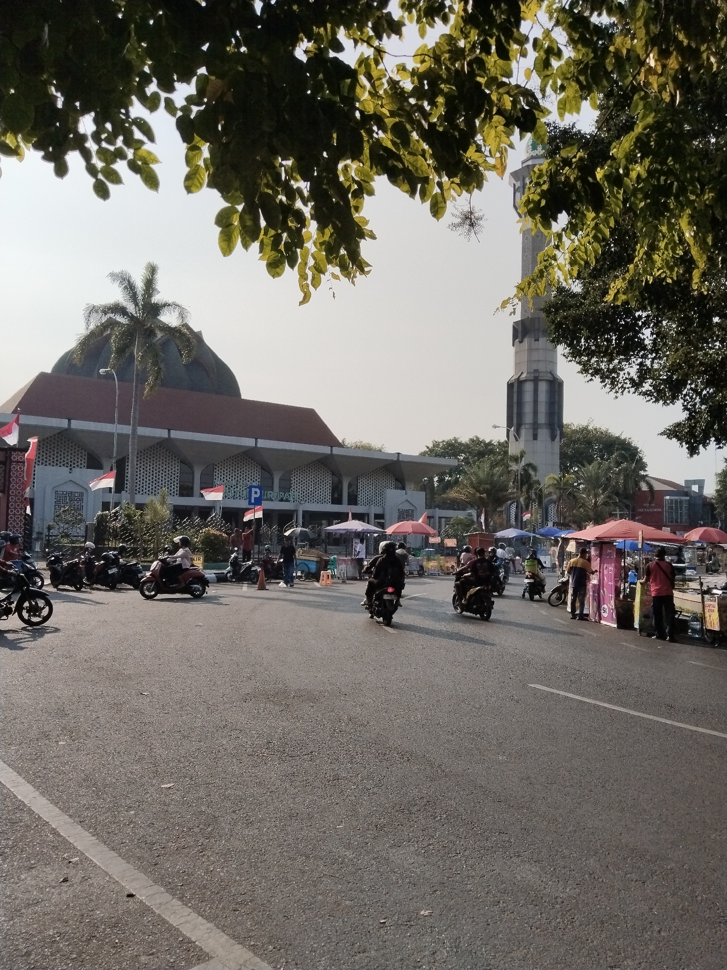 This image captures a street scene in Kecamatan Pati, Indonesia, featuring a prominent building that appears to be a mosque or a similar large public structure with a dome and a tall, multi-tiered minaret. The architecture is characterized by a white facade with repeating geometric patterns in the windows and a reddish-brown tiled roof. In front of the building, a row of street vendors' stalls with red and pink umbrellas lines the road, some displaying signage. Several individuals are visible near the stalls, some appearing to be vendors and others customers. The street itself is asphalt with a white lane marker. A number of motorcycles are parked and in motion on the road, with several riders visible. Indonesian flags are displayed on poles near the building. Lush green trees, including a palm tree, frame the upper portion of the image, suggesting an outdoor, potentially warm climate. The sky is overcast, and the lighting indicates it is daytime.