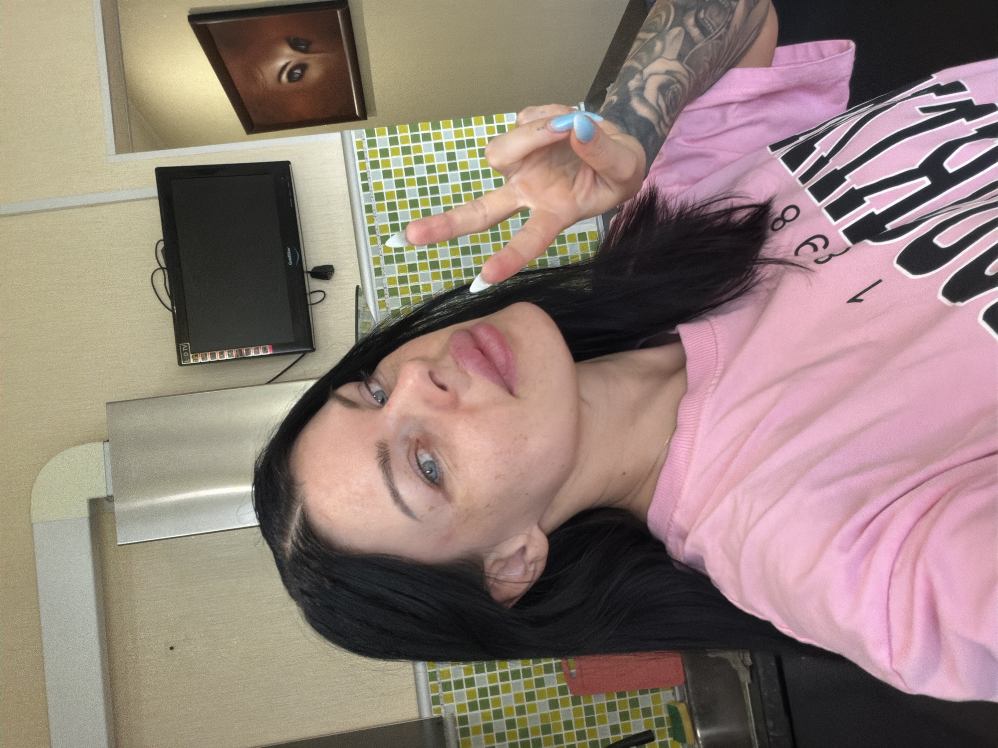 A woman with long, dark hair and blue eyes is taking a selfie in a kitchen. She is wearing a pink t-shirt with the word "BROOKLYN" printed on it and is making a peace sign with her right hand. She has light blue painted nails and a large tattoo on her left arm. The kitchen has light yellow walls with a checkered backsplash. A small television is mounted on the wall above a stainless steel exhaust fan. A framed picture of eyes is hanging on the wall to the right. The woman is positioned in the foreground, with the kitchen background slightly out of focus. The location is Ufa, Russia.