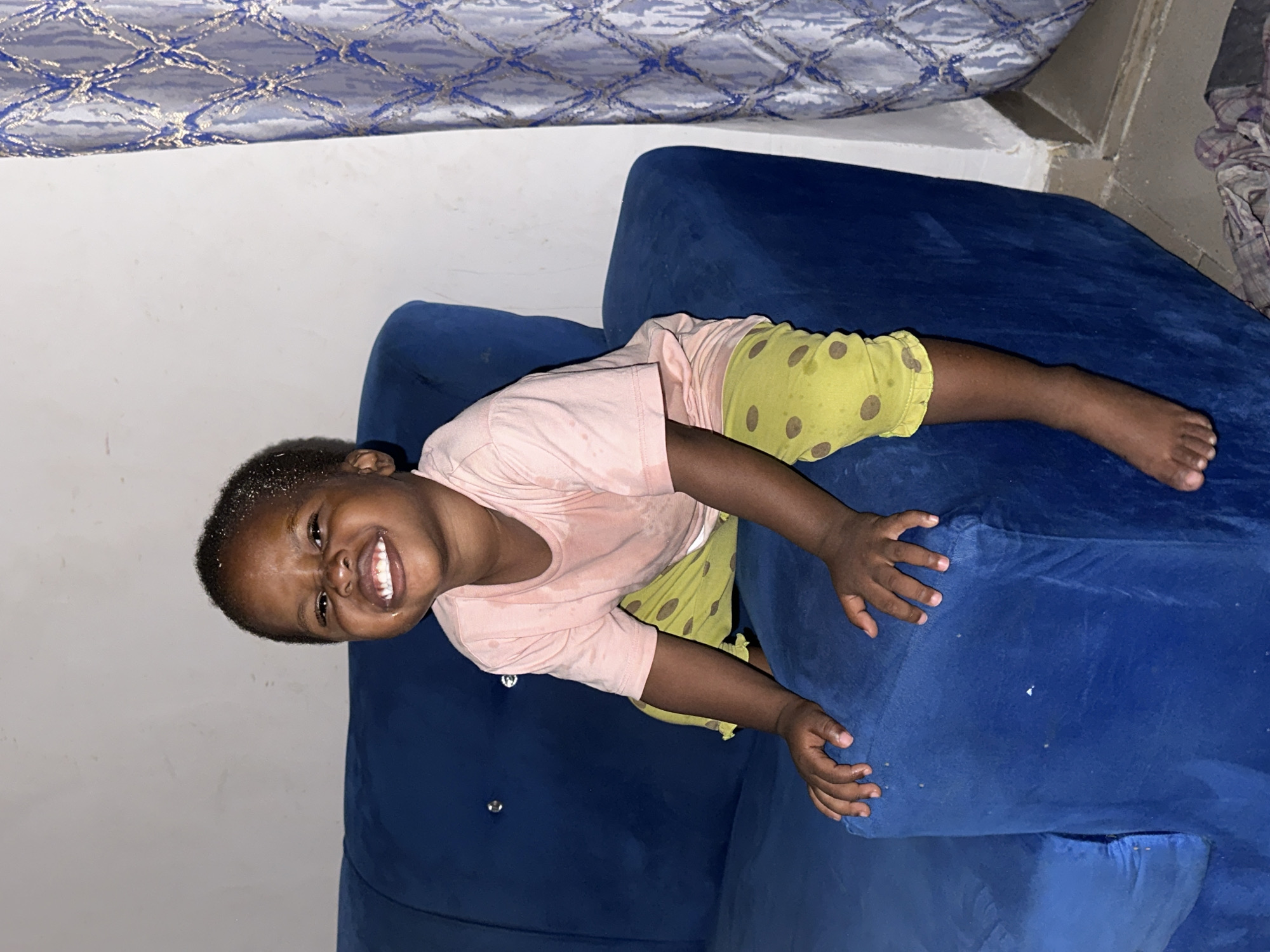 A young Black child is sitting on a blue sofa in what appears to be a room. The child is wearing a pink t-shirt and green pants with brown polka dots. The child is smiling widely, with their teeth showing. The child's hands are resting on the sofa in front of them. In the background, a white wall and a patterned curtain can be seen. The image is taken from a slightly high angle. There is no indication of the location being Narayi, Nigeria.