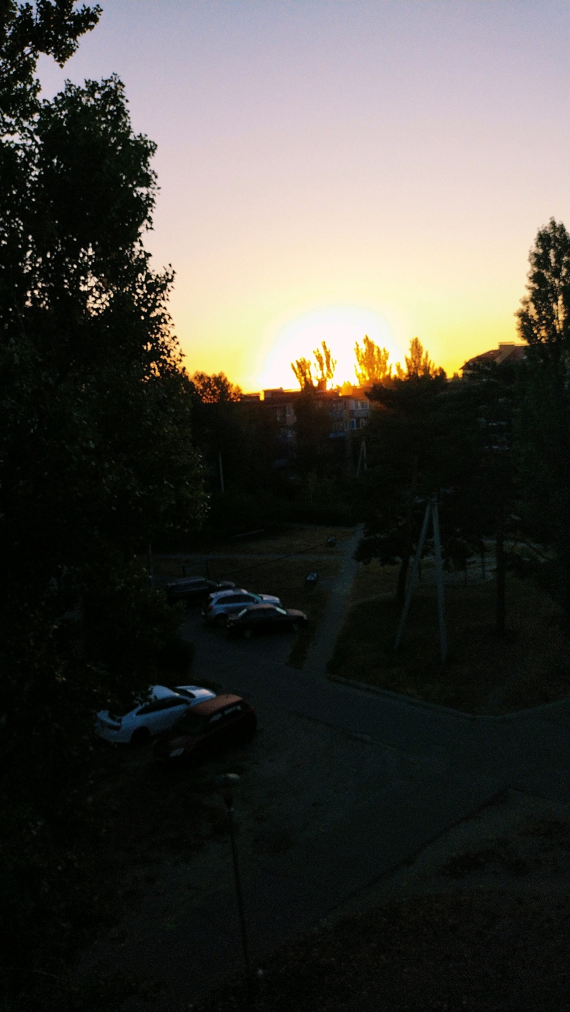 The image depicts a sunset over a residential area in Horishni Plavni, Ukraine. The sun, partially obscured by trees and buildings, casts a warm orange glow across the sky. In the foreground, silhouetted trees frame the scene, while a street with parked cars is visible below. The architecture suggests a typical Eastern European urban setting. The overall impression is one of a peaceful, everyday moment captured at dusk.