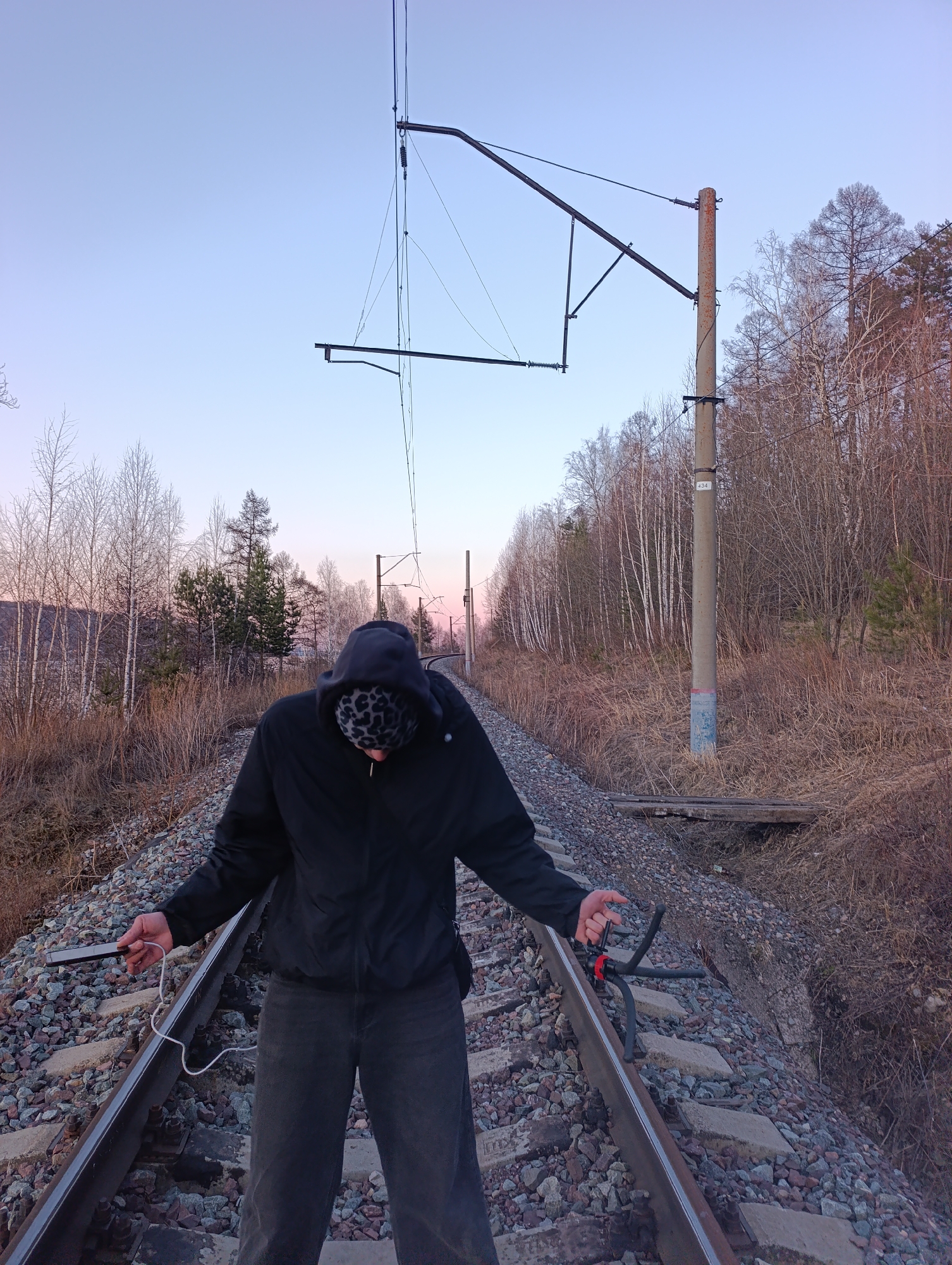 The image shows a person standing on a railroad track. The person is wearing a black hooded jacket and dark-colored pants. The hood is up and appears to have a patterned lining. They are holding a smartphone in their left hand, which is connected to a white cable. A black tripod is positioned on the right rail. The railroad track is surrounded by gravel and leads into a wooded area. Overhead electrical lines and support structures are visible above the track. In the background, there is a line of trees, primarily deciduous. The sky is a pale gradient of color, suggesting either sunrise or sunset. A utility pole labeled "434" is located along the tracks to the right of the frame. The image likely depicts a scene near the railroad tracks running through the outskirts or industrial areas near Krasnoyarsk, Russia, given the regional appearance of the trees and the power system infrastructure. The person may be taking photos or recording video using their smartphone and tripod.