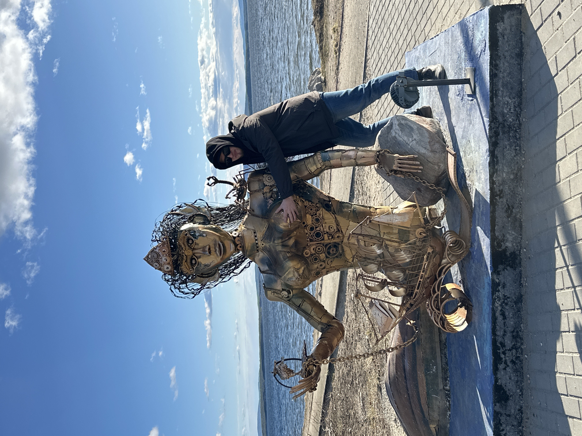 The image shows a man posing next to a metal sculpture of a mermaid. The man is wearing a dark hooded jacket, blue jeans, and dark shoes. He also appears to be wearing sunglasses. He is leaning on the sculpture with his right hand resting on the sculpture's chest. The sculpture is made of metal and depicts a mermaid with intricate details. The sculpture is placed on a concrete base, which is painted blue. In the background, there is a body of water with visible shoreline, and a blue sky with scattered clouds. The scene is set on a paved area, possibly a walkway or promenade. The location is in the city of Unknown, Unknown.