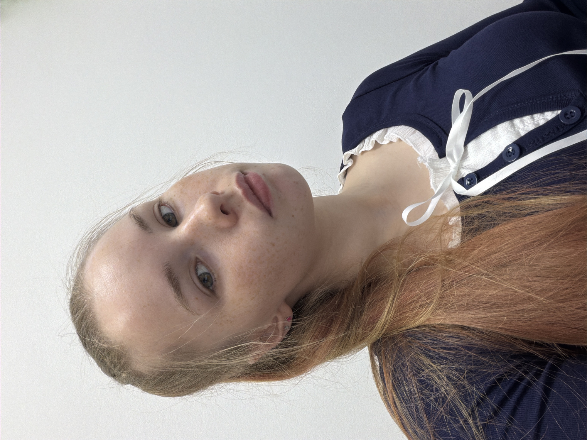 A close-up, tilted portrait shows a fair-skinned young woman with freckles. She has light-colored eyes and reddish-blonde hair partially pulled back. She is wearing a dark blue top with white trim and a white ribbon tie at the neckline. The background is a plain, white surface. There is no information to determine the location in Berezniki, Russia.