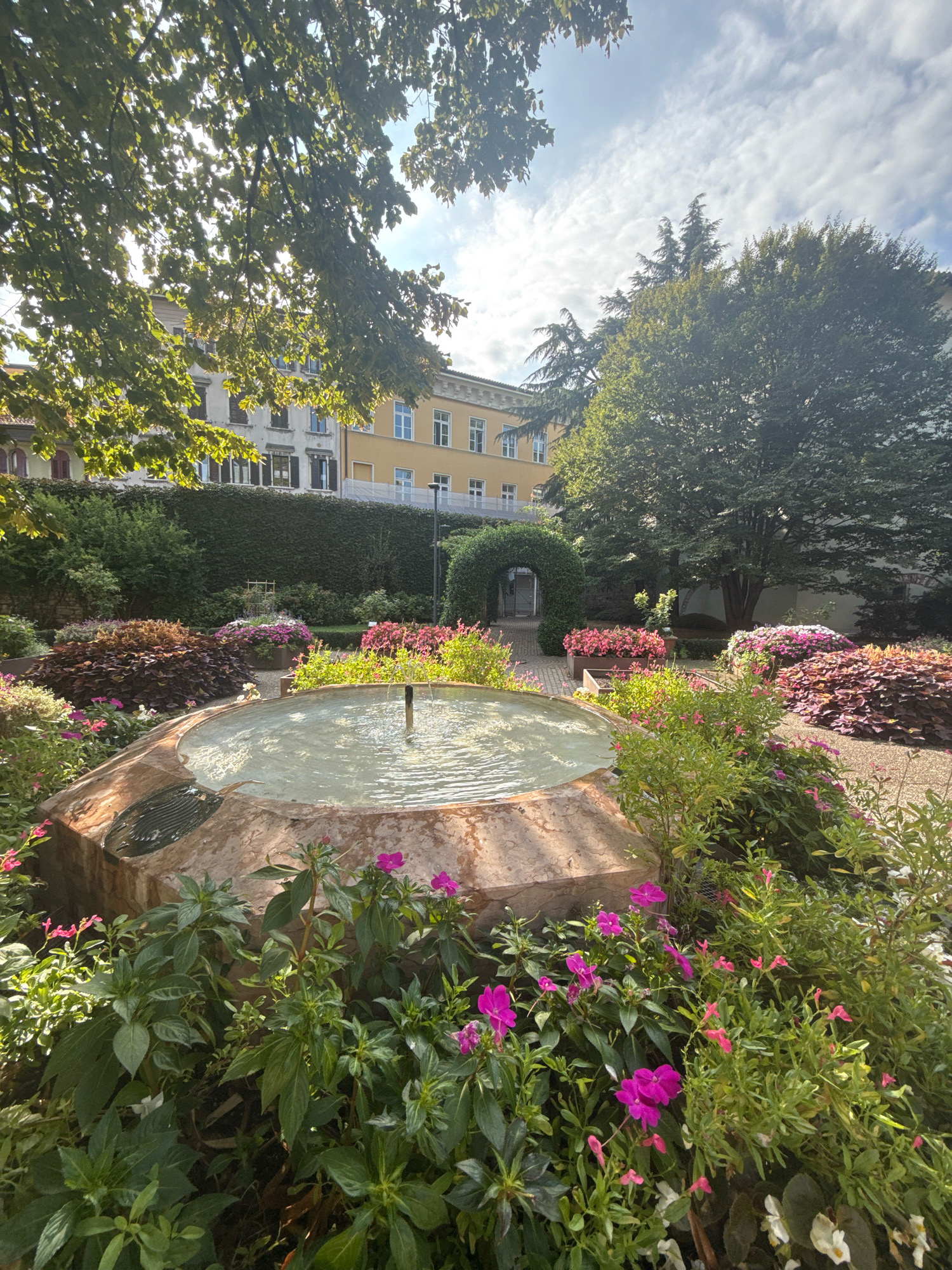 The image depicts a tranquil garden scene in Trento, Italy. In the foreground, a large, octagonal stone fountain with a light-colored marble basin is filled with clear water, with a dark, circular insert visible on the left side of the basin. Water gently spills over the edge of the fountain. The fountain is surrounded by lush greenery and vibrant pink and purple flowers. Behind the fountain, a path leads to a meticulously maintained hedge archway, suggesting an entrance to another area of the garden. Beyond the archway, large, mature trees stand tall, their branches casting dappled shadows. In the background, a building with a pale yellow facade and white-framed windows is visible, partially obscured by trees. The sky above is a mix of blue and white clouds, with sunlight filtering through the leaves of a large tree in the upper left corner of the frame. The overall impression is one of a peaceful, well-kept urban garden.