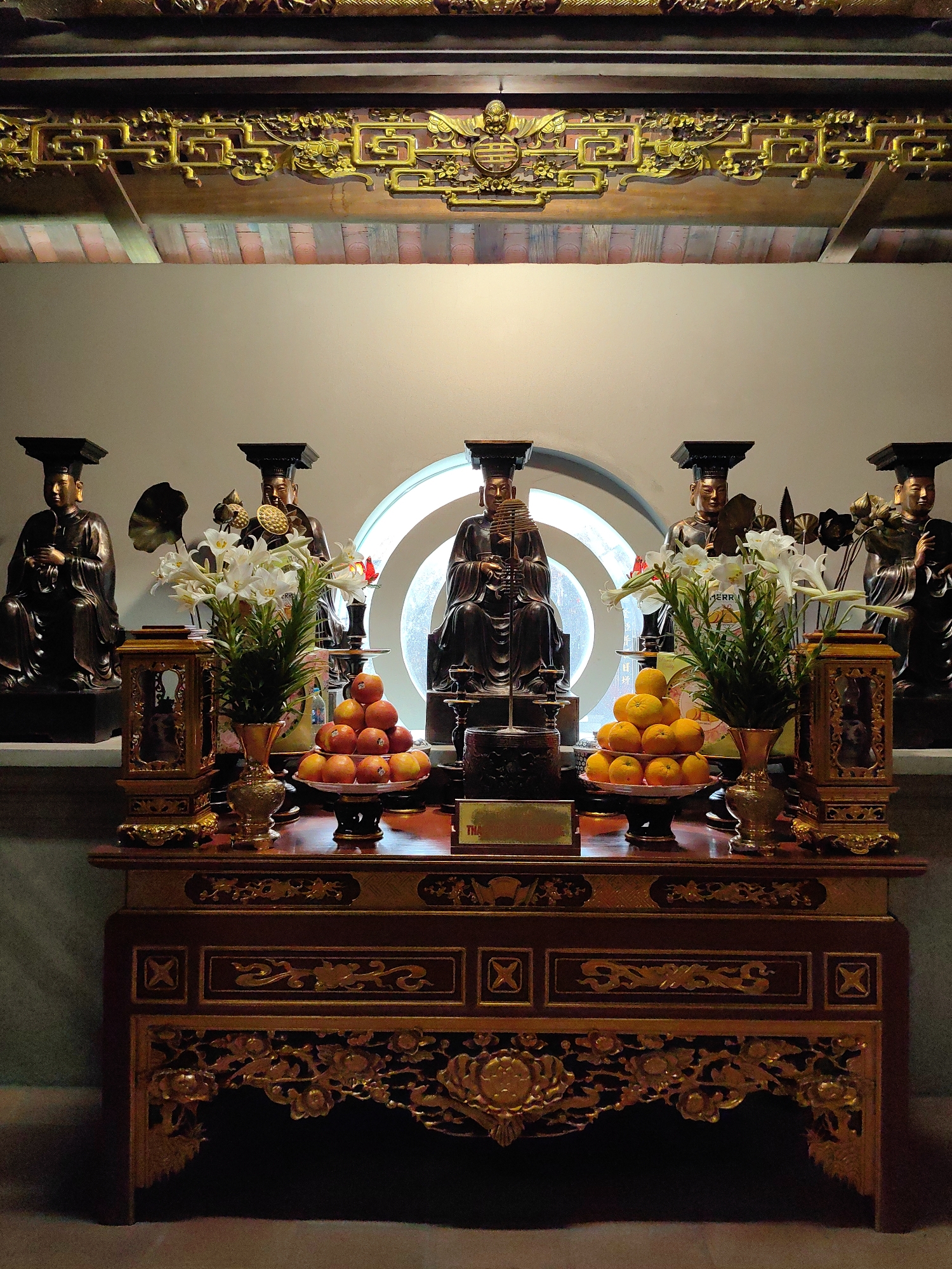 In Hong Gai Ward, Vietnam, an altar is positioned in front of a circular window. Statues of seated figures wearing rectangular hats sit on a shelf behind the altar. The altar is adorned with two pyramids of fruit on plates and vases of white lilies. Ornate, golden trim decorates the edges of the altar and a beam overhead.