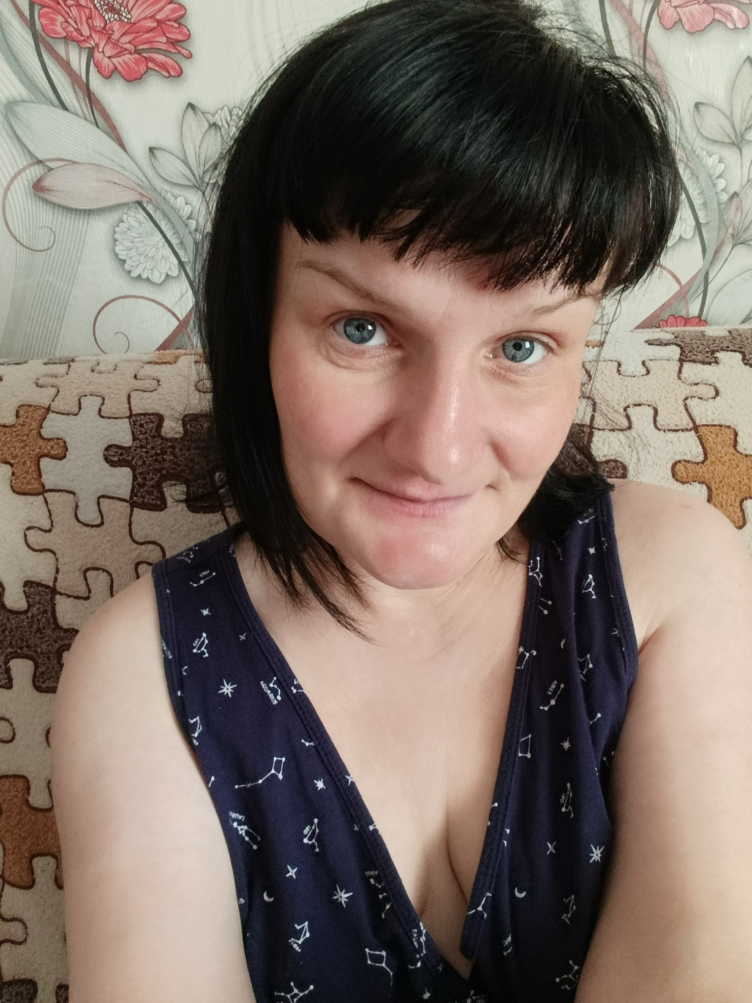 A head-and-shoulders portrait shows a woman looking directly at the camera with a slight smile. She has short, dark, chin-length hair with bangs and striking blue eyes. Her skin appears fair. She is wearing a dark blue V-neck sleeveless top adorned with white patterns of constellations, stars, and crescent moons. Visible text on her top includes "LEO," "AQUARIUS," and "ARIES."
Behind her, a light-colored couch or cushion with a brown and beige puzzle-piece pattern is visible. Further in the background, a wall is covered with wallpaper featuring a floral design. This design includes prominent red and pink flowers, along with lighter grey and white floral and leaf motifs. The overall setting appears to be indoors. The lighting is soft and even, suggesting an indoor environment without strong direct sunlight. The woman's expression is calm and pleasant.