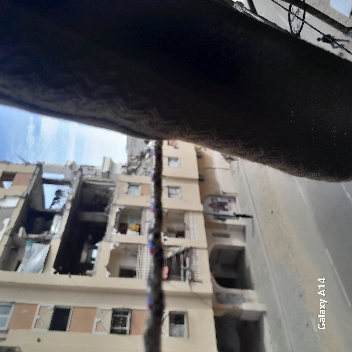 The image depicts an urban street scene in Khan Yunis, Palestinian Territory, captured from an elevated perspective. In the foreground, a dark, textured fabric object partially obstructs the right side of the frame, and a horizontal rope with colorful segments (red, blue, white) stretches across the center.
The background features a multi-story, light-colored building exhibiting significant structural damage, particularly on its upper floors, where large sections of exterior walls are missing, exposing interior spaces and debris. Rebar and concrete rubble are visible within these damaged areas. Undamaged lower floors include balconies, with clothes visible hanging on one.
Below the buildings, a paved street with a double white line marking is visible. Two individuals in dark clothing are present on or near the street. One person walks on the sidewalk in the mid-left portion of the frame, while another is partially visible in the bottom-right. The sky above is partly cloudy. The image was captured with a "Galaxy A14."