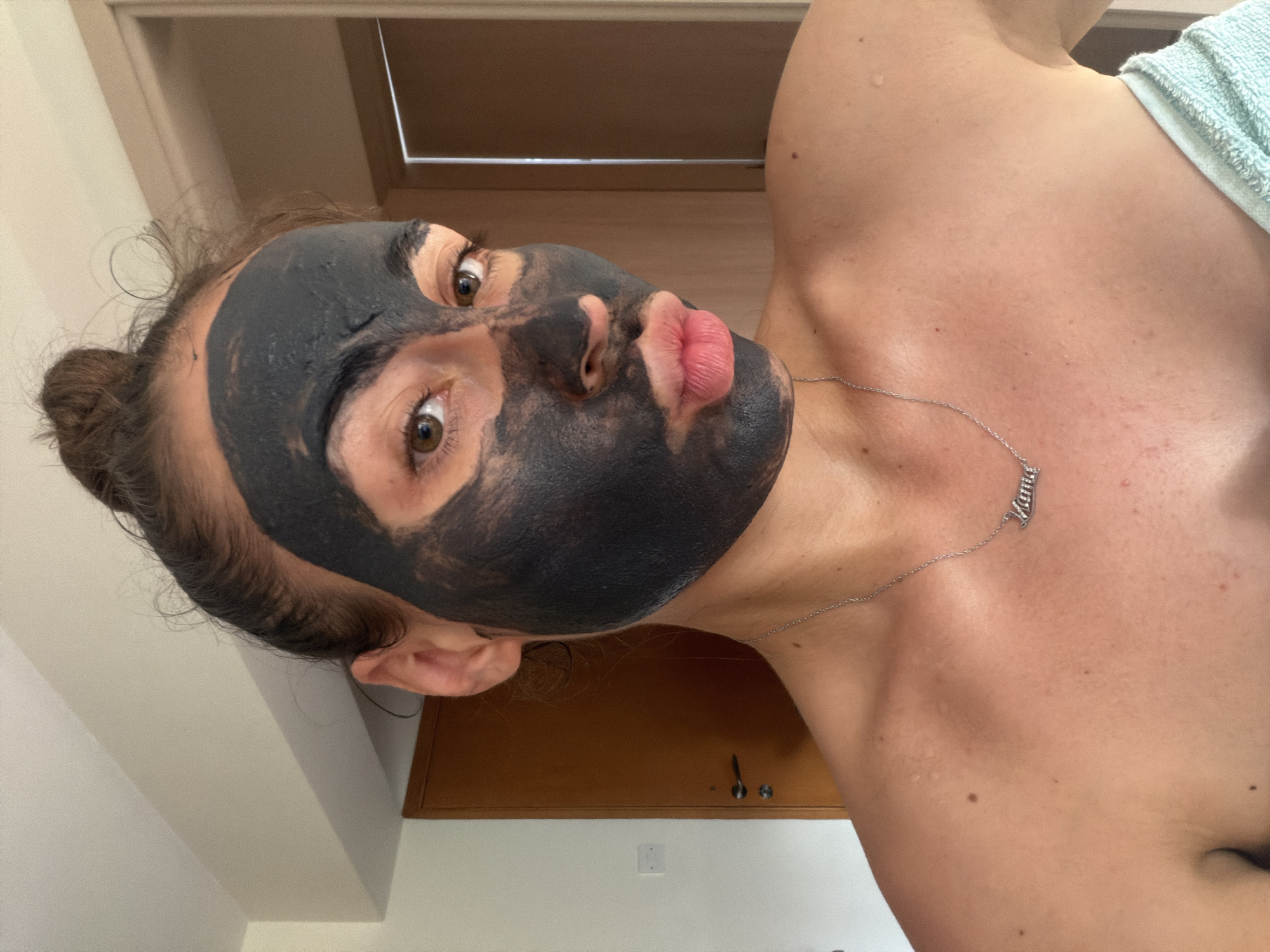 A young woman with a dark mud mask on her face is pictured from the chest up in an indoor setting, likely a home bathroom. She is looking directly at the camera with her lips pursed. A silver necklace with the word "Mama" is visible around her neck. Her hair is pulled up in a bun. The background includes a closed wooden door and a white wall with a light switch. The lighting suggests it is daytime.