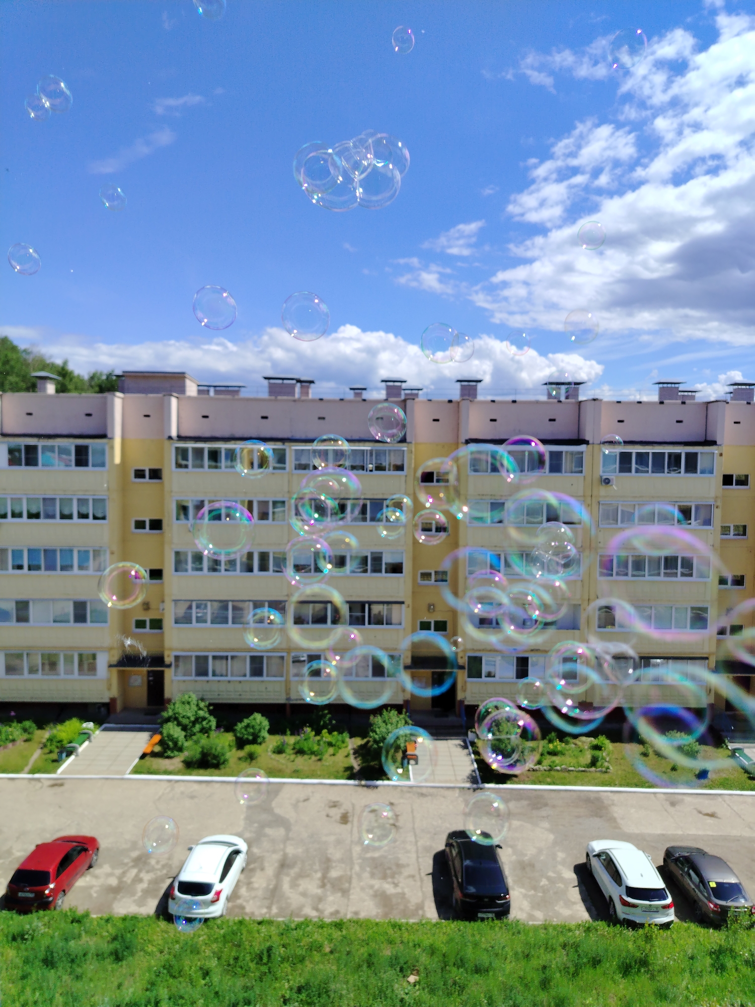 An elevated daytime view depicts a residential area in Noviy, Russia, under a bright blue sky with scattered white cumulus clouds. Numerous iridescent soap bubbles of varying sizes float through the air, displaying rainbow refractions across the frame, predominantly in the mid-ground. The central feature is a multi-story residential building with a light yellow facade and a pale brown upper section, punctuated by numerous white-framed windows and enclosed balconies. Green foliage, including trees and bushes, is visible around the building's foundation. A light grey paved area, likely a parking lot, lies below the building, where five cars are parked in a row facing left: a red hatchback, a white hatchback, a black sedan/SUV, a second white sedan/SUV, and a dark grey sedan/SUV. A vibrant green grassy strip occupies the immediate foreground. No people are present.