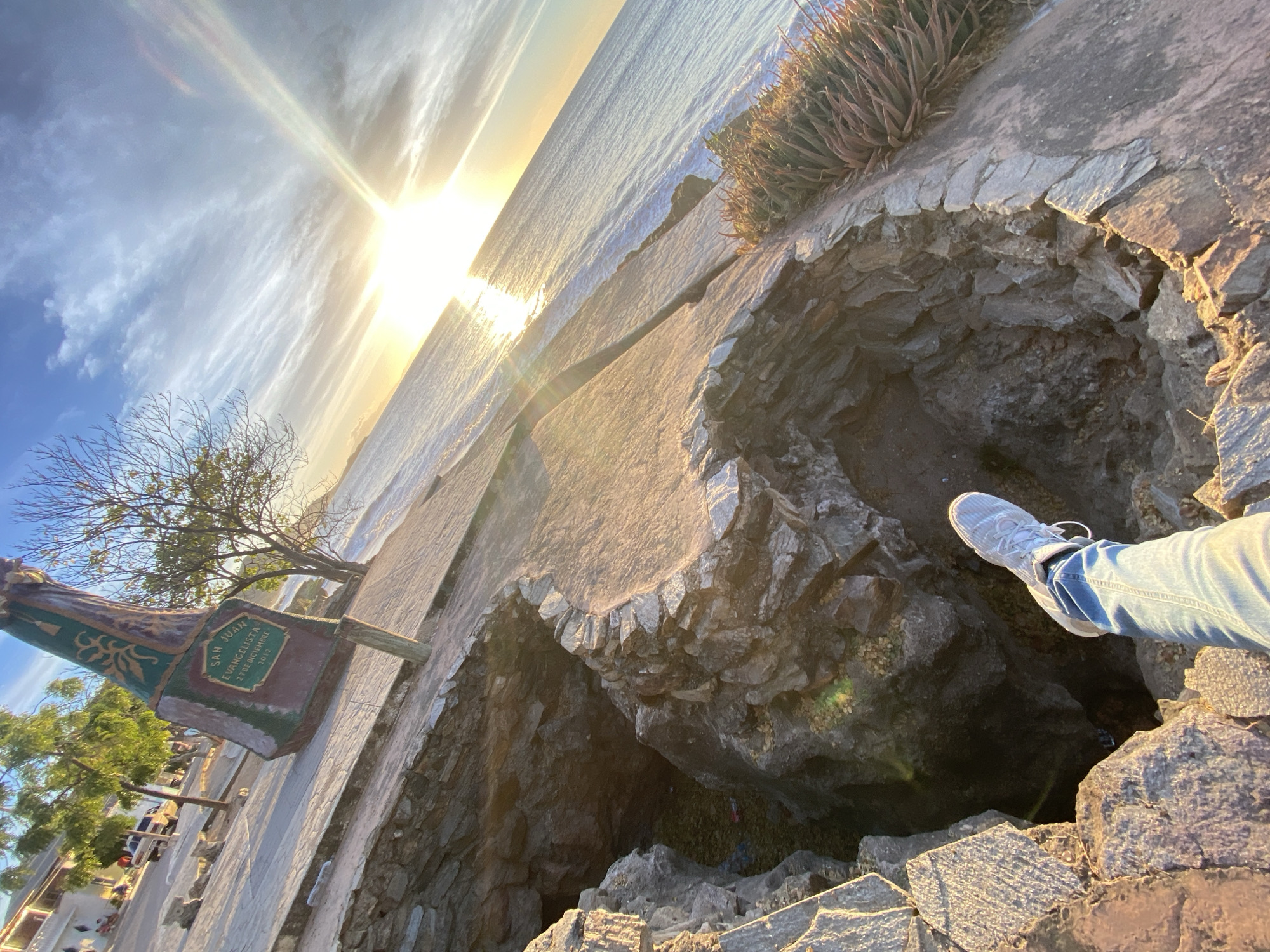 A low-angle shot oriented sideways depicts a cliffside scene in Juan Griego, Venezuela. A person's leg and sneaker extend into a natural rock cavity or hole near the cliff edge. The ocean is visible in the background under a bright, sunny sky. A statue bearing the inscription "San Juan Evangelista, 27 de Diciembre 2012" is placed on the walkway to the left. The surrounding environment consists of stone structures, sparse vegetation, and a coastal walkway leading to a town with several buildings and vehicles.