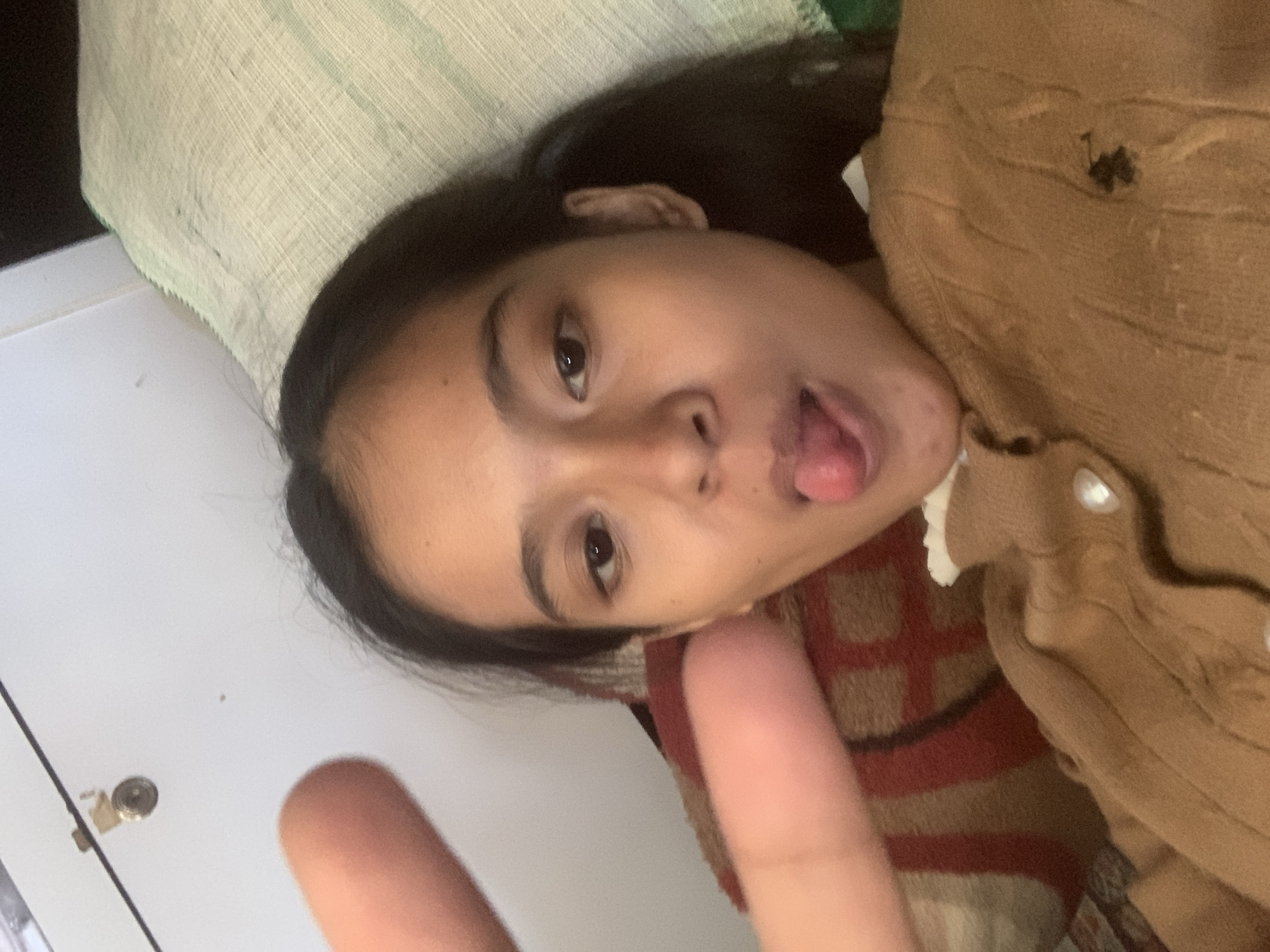 In Sungai Meriam, Indonesia, a young female is captured in a close-up, selfie-style photograph. She has dark hair and is wearing a tan cable-knit cardigan with white buttons. Her face is turned towards the camera, and she is sticking out her tongue playfully. Her left hand is visible in the foreground, held up in a peace sign. She is lying down, as indicated by the presence of a pillow with a light green cover and a patterned blanket behind her. The background includes a white wall with a small metallic fixture and a dark element that could be furniture or a wall. The lighting suggests an indoor environment.