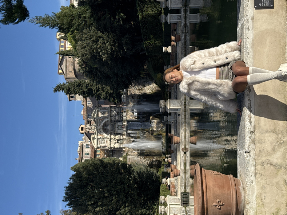 The image depicts a serene outdoor scene at the Villa d'Este in Tivoli, Italy, under a clear blue sky. In the background, the hillside town of Tivoli is visible, characterized by buildings with terracotta roofs and a prominent round tower structure, framed by dense green trees. Below the town, a grand Renaissance-era fountain complex, consistent with the Organ Fountain or Neptune's Fountain at Villa d'Este, dominates the mid-ground. This elaborate structure features multiple tiers, architectural embellishments, and several powerful jets of water cascading into pools below.
In the mid-ground and extending into the foreground, a large, rectangular reflecting pool lies still, mirroring the sky and surrounding architecture. Numerous empty terracotta planters of various sizes are arranged symmetrically along the edges of the pool.
In the immediate foreground, positioned on the lower right, a person with medium-length brown hair, appearing to be of East Asian descent, is seated on a low, weathered stone ledge. The individual is wearing a light cream-colored faux fur coat over a white ribbed turtleneck top, a plaid mini-skirt with beige, brown, and black tones, light grey knee-high socks, and white sneakers. Their eyes are closed, head slightly tilted upward, and a subtle smile is present, suggesting repose or enjoyment of the sunlight. The strong sunlight casts a distinct shadow of the person onto the stone wall and ground to their right. A large terracotta planter with a fleur-de-lis emblem is visible in the bottom left corner, and a small, dark informational sign with white text is affixed to the ledge to the right of the seated individual.