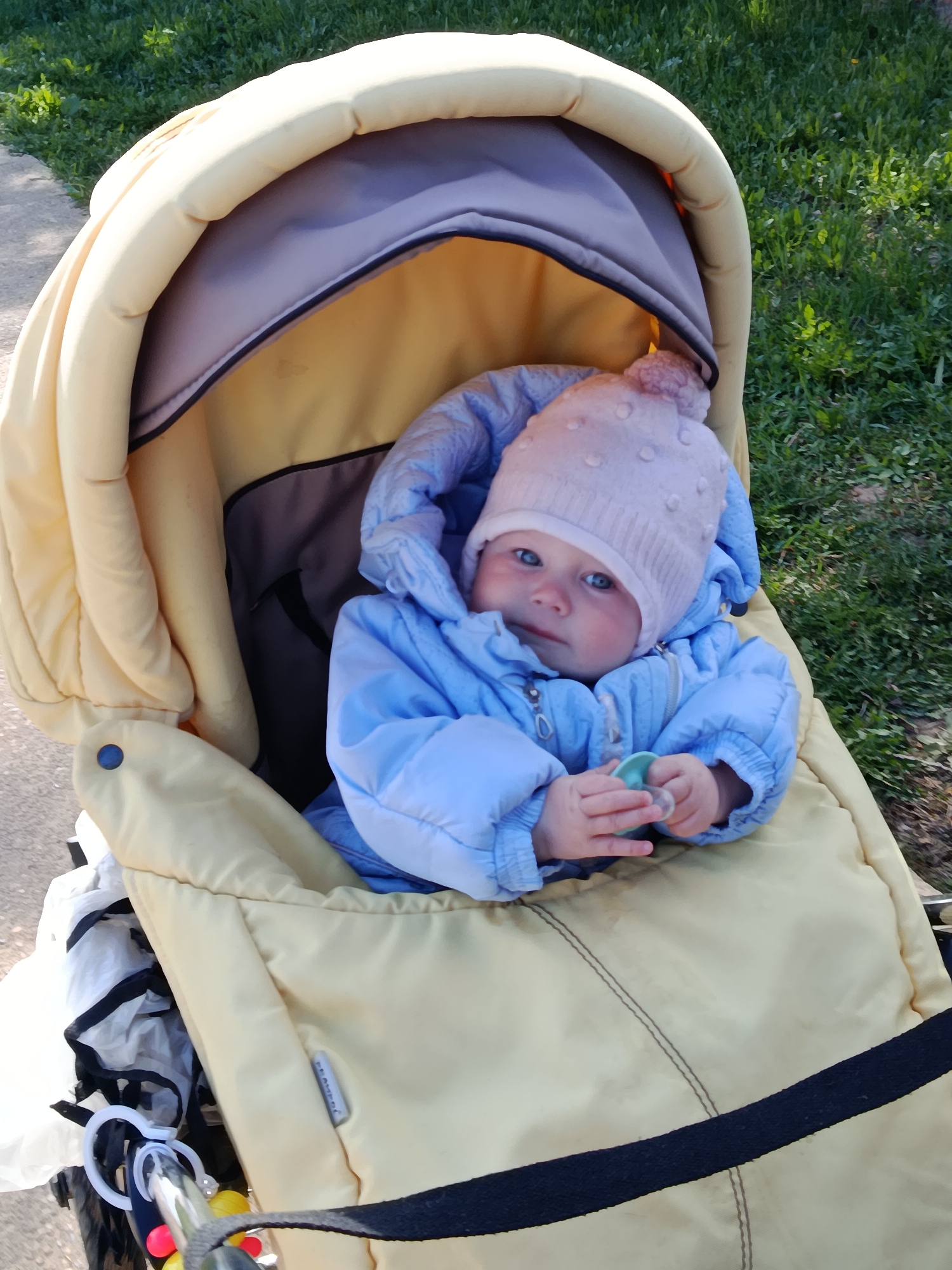 A baby dressed in a light blue jacket and a pink knit hat with polka dots sits in a tan stroller with a raised canopy. The baby is holding a light green pacifier. The stroller is parked on a sidewalk with grass visible in the background. The location is in the city of Фоки, Russia.