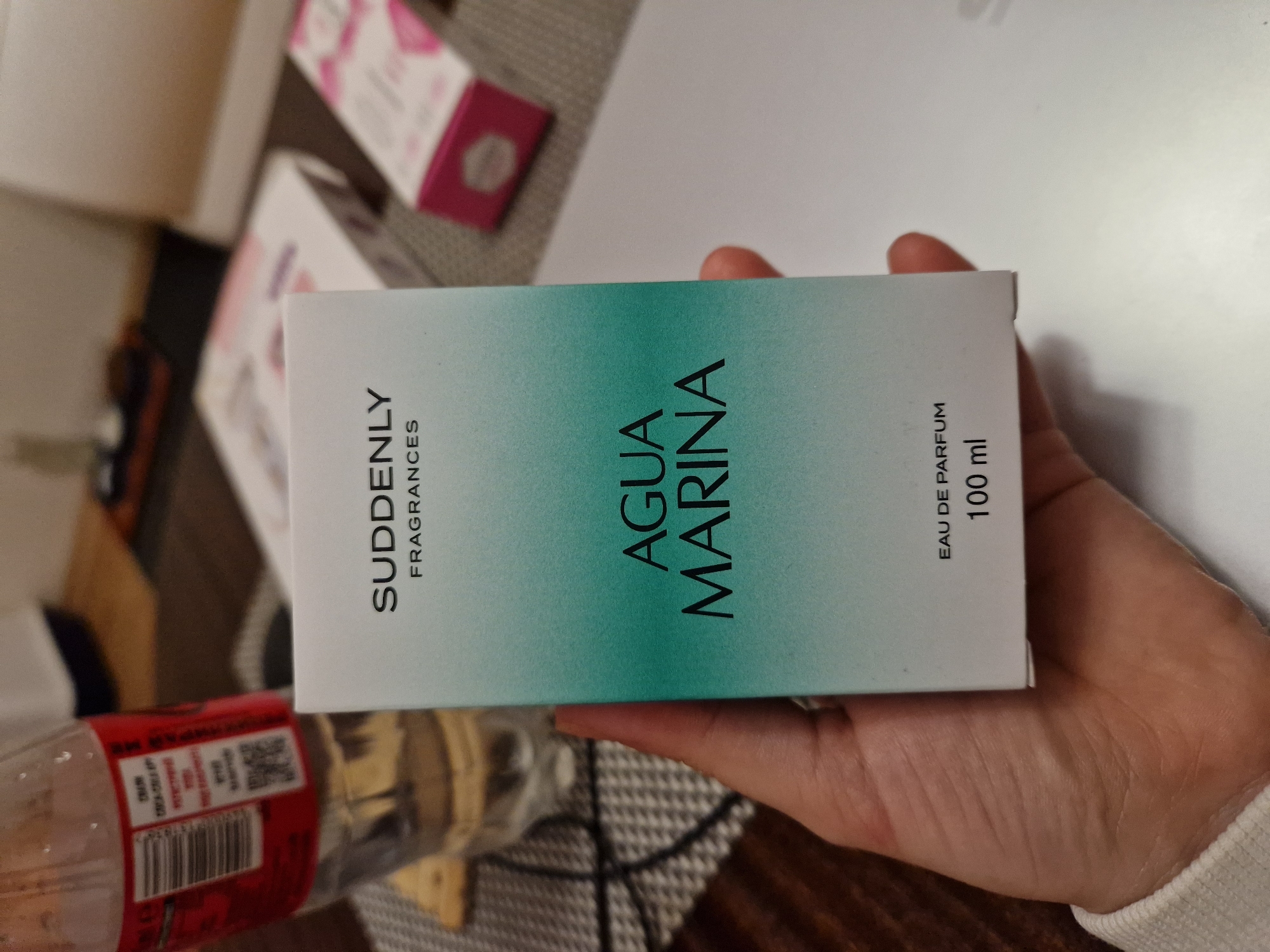 A hand holds a box of "Suddenly Fragrances Agua Marina Eau de Parfum 100 ml". Other boxes and a water bottle are in the background. The location is unknown, and there is no explicit information to connect it to Lovech, Bulgaria.