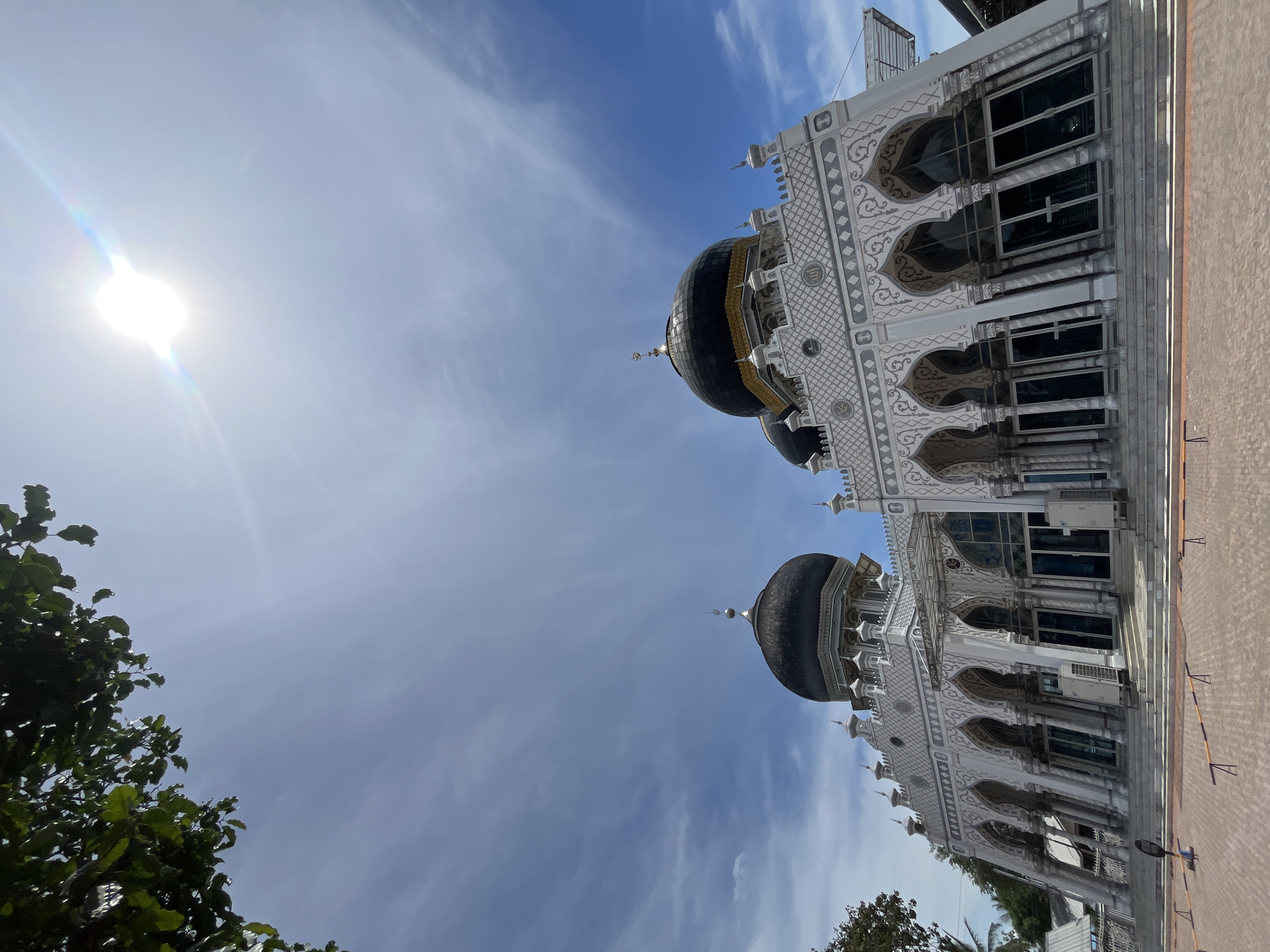 The image shows a mosque against a sky with a bright sun on the left and streaky clouds. The mosque appears to be white with darker domes and detailed archways. Part of a tree is visible at the bottom left corner. The image is taken from a low angle and appears to be rotated. The city of Keudekruenggeukueh, Indonesia, is given as the location.