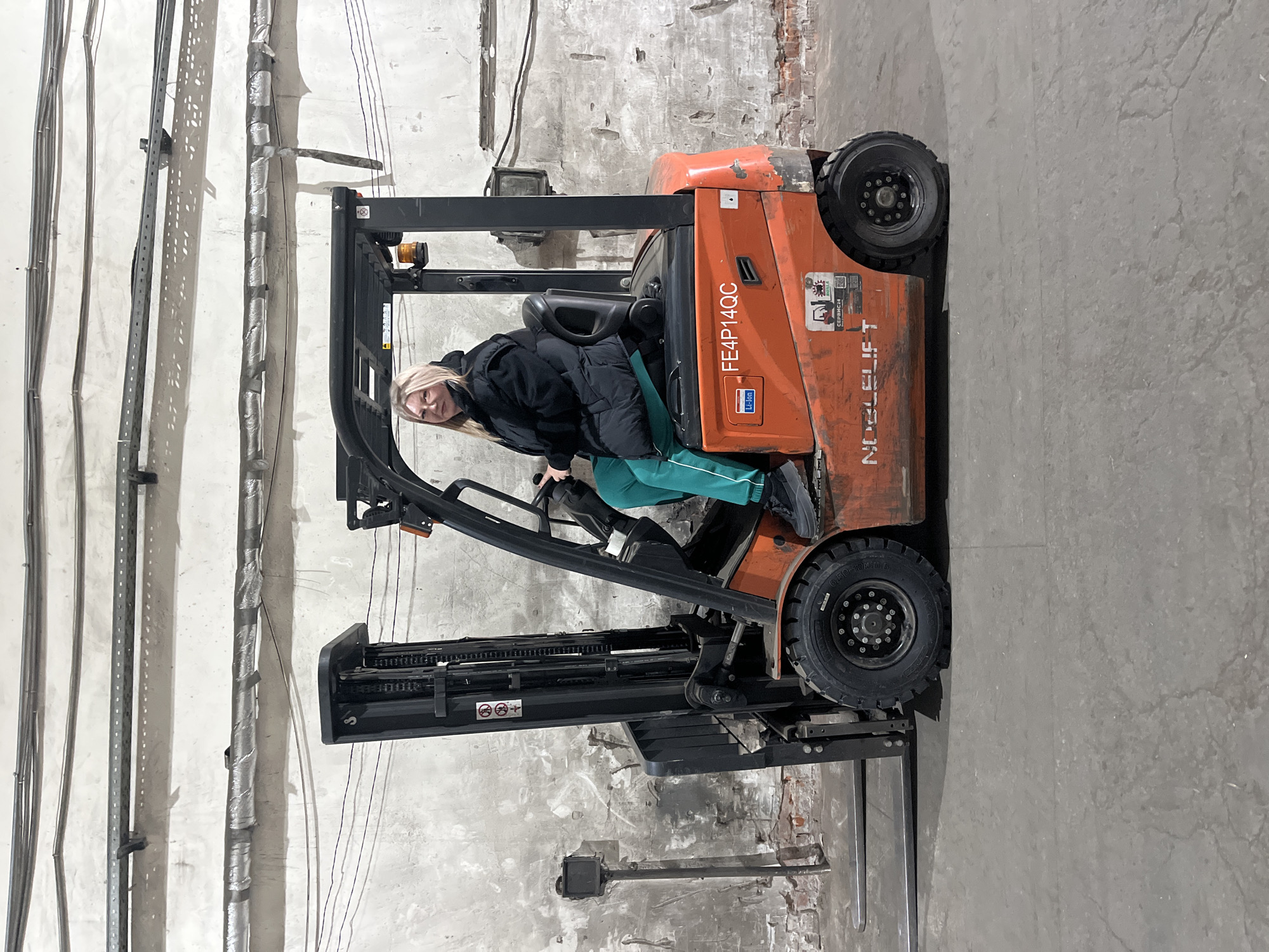 A blonde female is seated in the operator's compartment of an orange NOBLELIFT FE4P14QC electric forklift. She wears a black puffer jacket, dark green sweatpants with a white side stripe, and black athletic shoes, looking towards the viewer with a slight smile. Her hands are positioned on the steering wheel. The forklift's black forks are extended forward, resting on the gray concrete floor. The vehicle's side features "Li-ion" and "СЕРВИС24" stickers, along with signs of wear and scuffing. The background wall is a worn, light-colored surface with visible plaster damage, exposed brick, and dirt. Numerous conduits, insulated pipes, and electrical wiring are affixed horizontally to the wall, along with a perforated metal cable tray. A vertical metal post topped with a small square component stands against the wall on the left. The scene depicts an indoor industrial or warehouse environment. The precise location within Izhevsk, Russia, cannot be determined from the visual evidence provided.