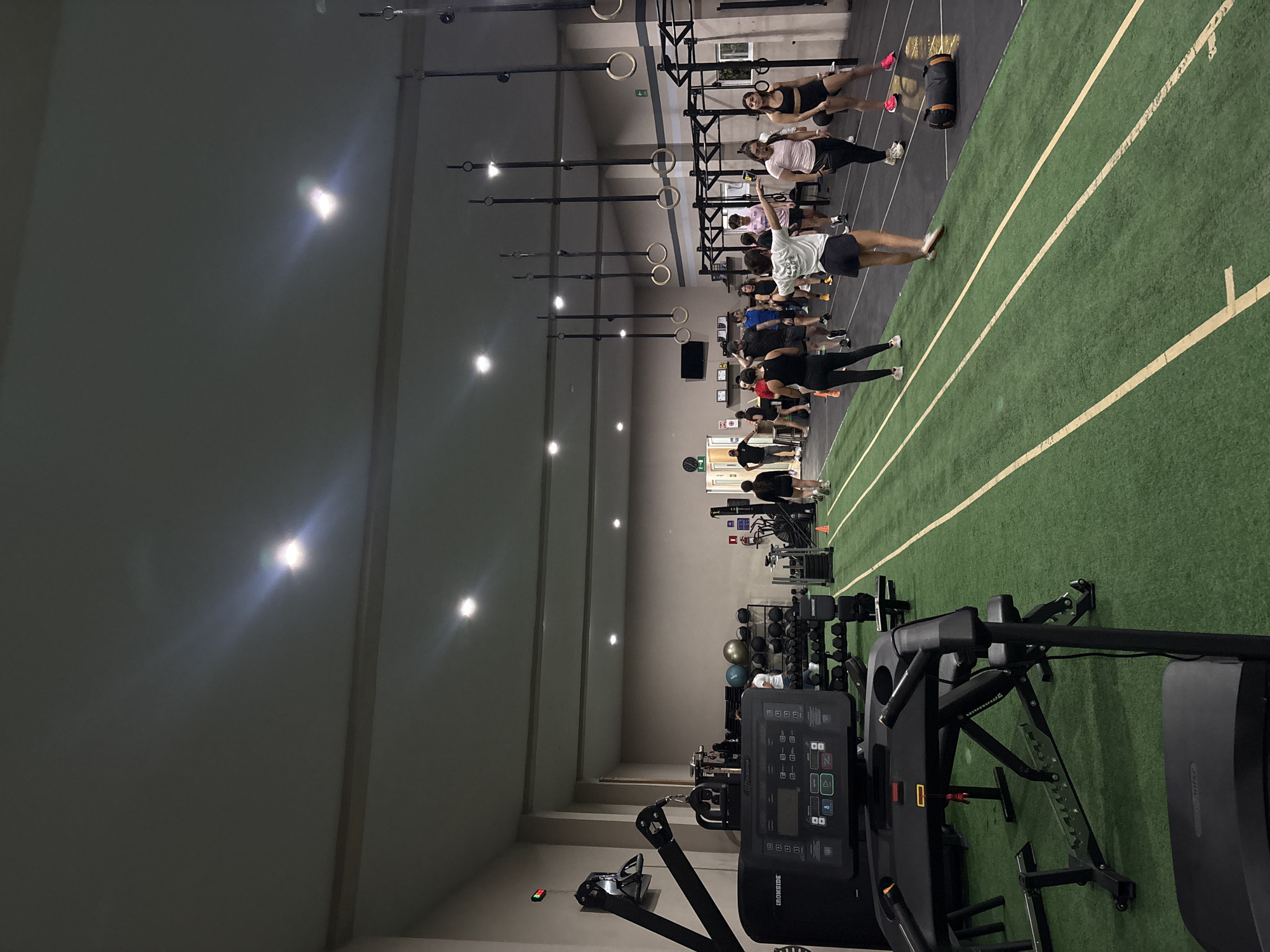 The image depicts the interior of a gym or fitness center in San Bernardino Tlaxcalancingo, Mexico. A group of people is engaged in a fitness class, standing in lines on a green artificial turf area marked with white lines. Overhead, gymnastic rings are suspended from a ceiling structure. In the foreground, various pieces of exercise equipment are visible, including a treadmill-like machine and a rack with weights. The room is well-lit by ceiling lights.