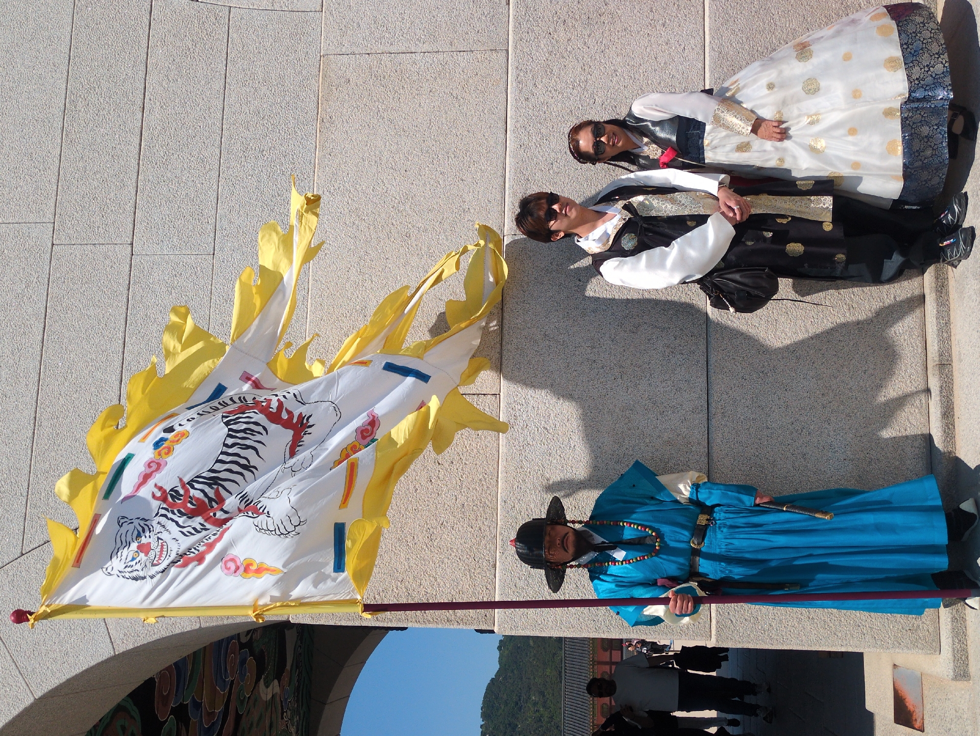The image, set in Seoul, South Korea, shows three individuals dressed in traditional attire next to a large flag with a tiger illustration. Two of them, a woman and a person are wearing traditional Korean Hanbok outfits. The third person is dressed in a blue traditional robe and hat. The flag they stand beside is adorned with a white background, a tiger drawing, and yellow flames along its edges. In the background, the top of a gate and a glimpse of a mountain can be seen. The scene is outdoors, likely in a historic or cultural site, possibly a palace or temple grounds. Other people can be seen in the background.