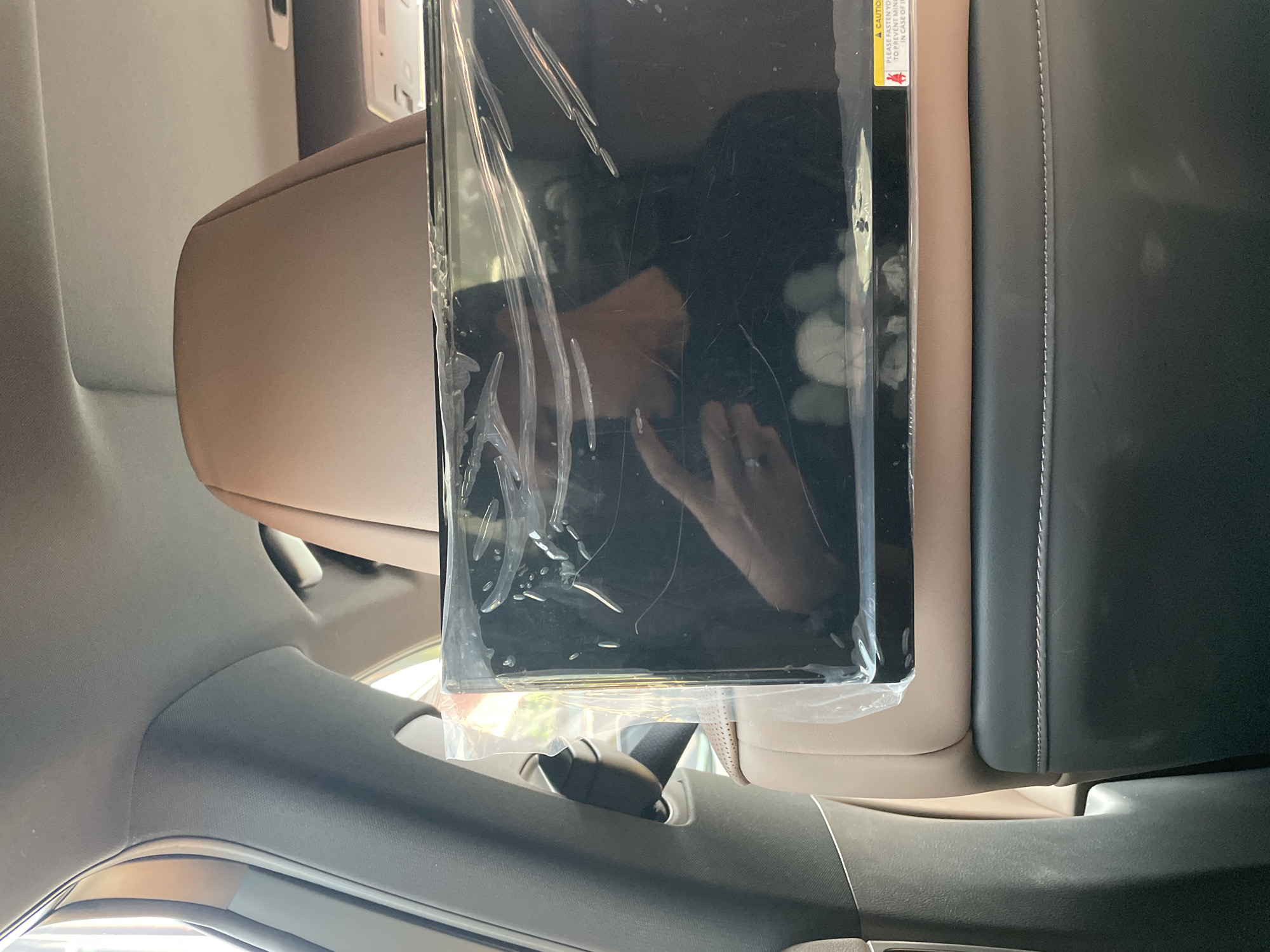 The image displays the interior of a vehicle in Ciater, Indonesia, viewed from a rear passenger seat looking forward. A dark-colored flat screen, encased in clear, wrinkled plastic protective wrapping, is affixed to the back of the front passenger seat's light brown leather headrest. The screen's reflective surface shows a clear image of the person taking the photograph, visible as a dark-clothed individual holding a device with their right hand, which wears a ring on the ring finger. A portion of the individual's face, including the nose and mouth area, is discernible in the reflection. Below the screen, the front seat's lower backrest is upholstered in a dark material featuring a single line of white horizontal stitching. A small yellow and red caution sticker, partially visible beneath the plastic wrap in the bottom right corner of the screen, displays text that begins "A CAUTION PLEASE FASTEN YO..." The vehicle's interior includes a dark grey pillar on the left and a light grey headliner above. Ambient daylight is visible through the left side of the car, illuminating parts of the interior.